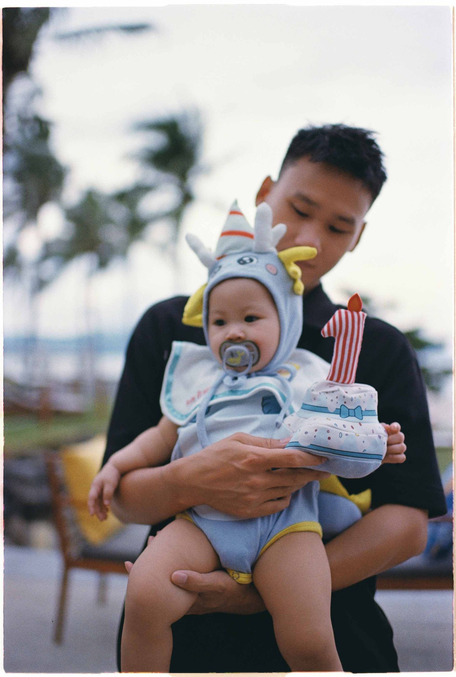 Da Nang family: baby wearing a cute animal-themed outfit and pacifier is held at a beachside location, by AnhVuWedding.