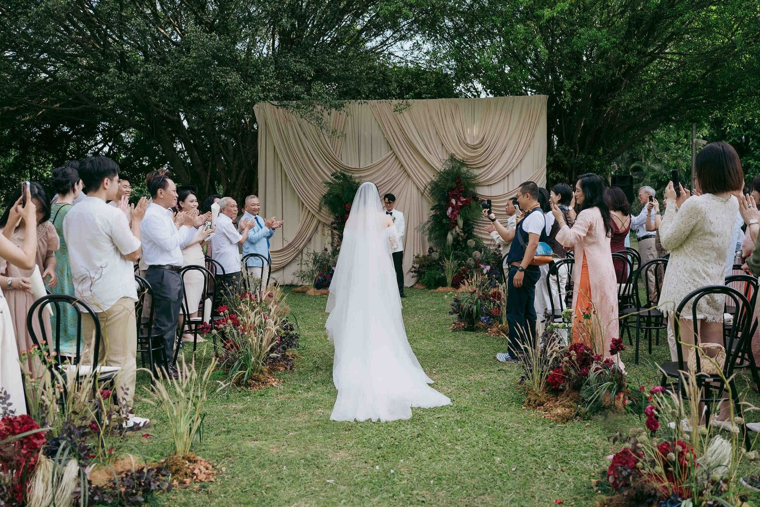 Outdoor garden wedding ceremony with bride in white gown and veil walking down aisle surrounded by guests and floral decor.