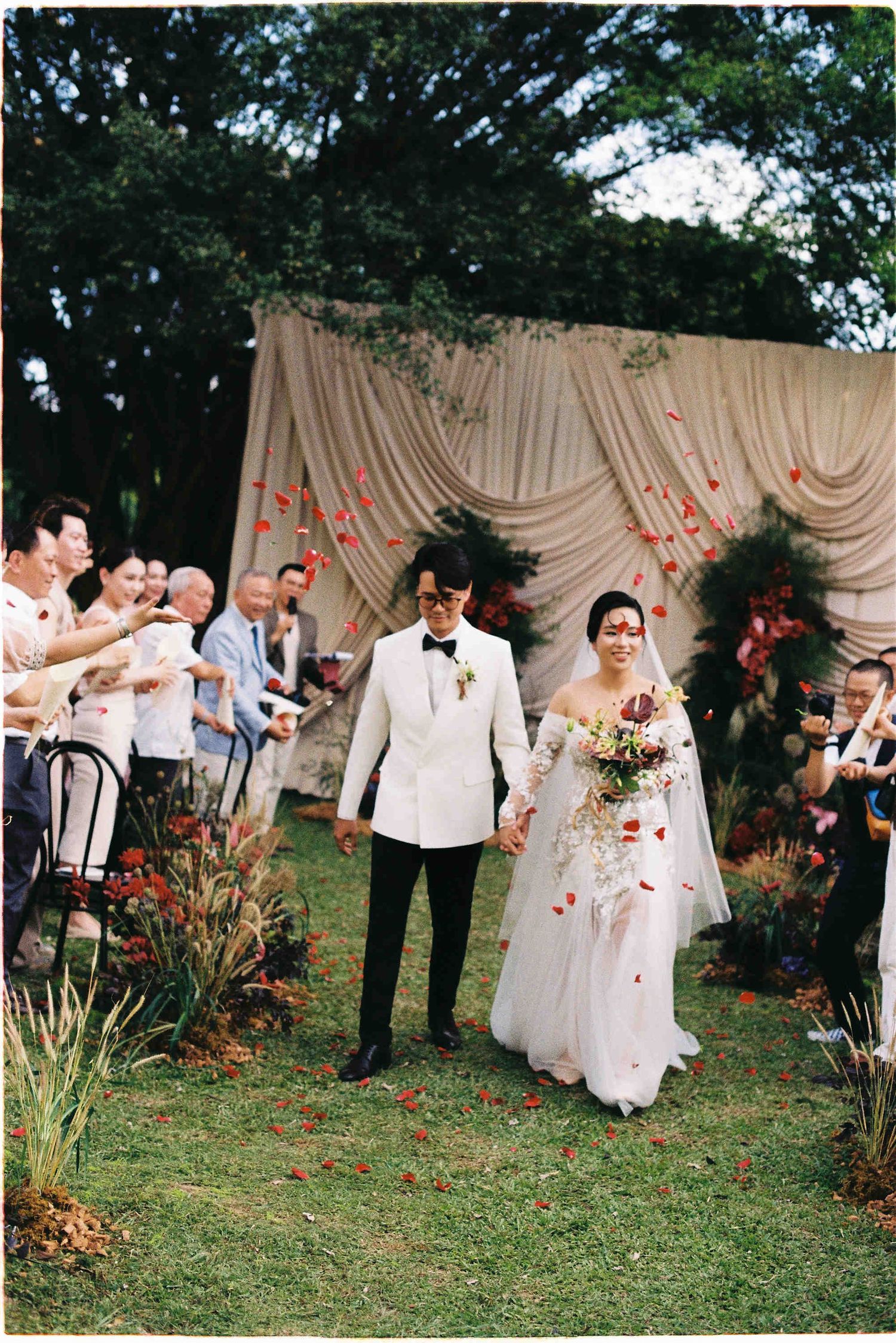 Newlywed couple walks down outdoor aisle as guests toss red rose petals, with draped fabric backdrop.
