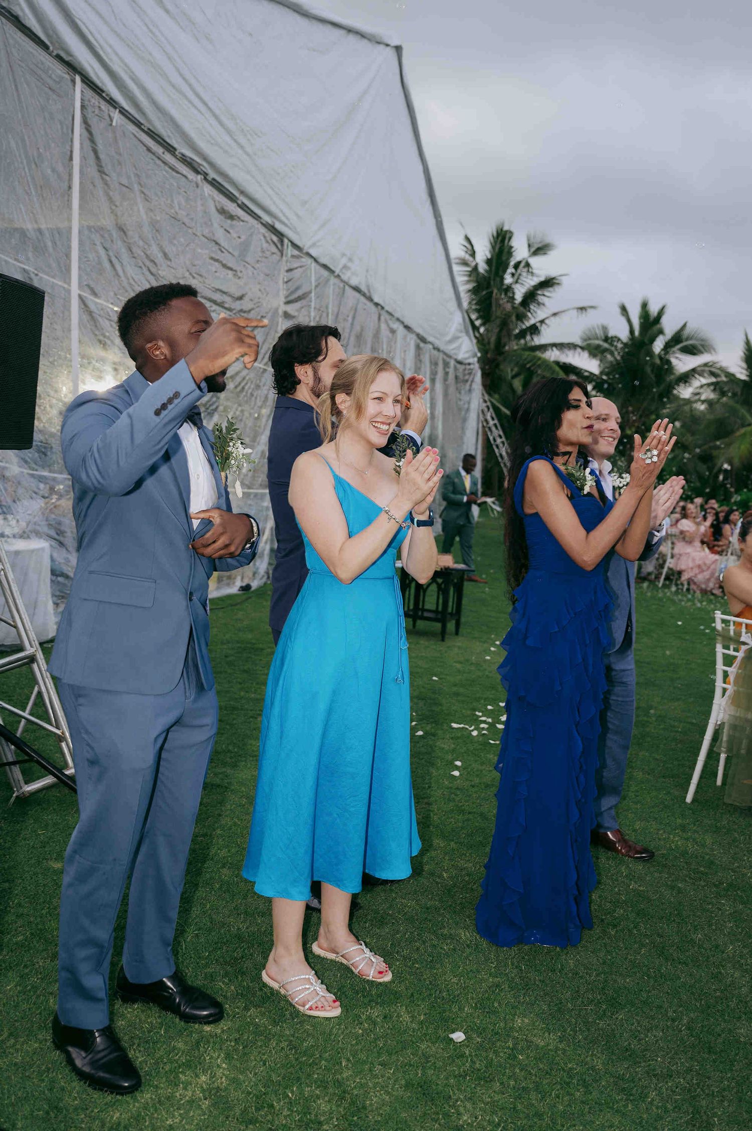 Wedding guests in blue attire clapping and celebrating outdoors near a white tent venue with palm trees.