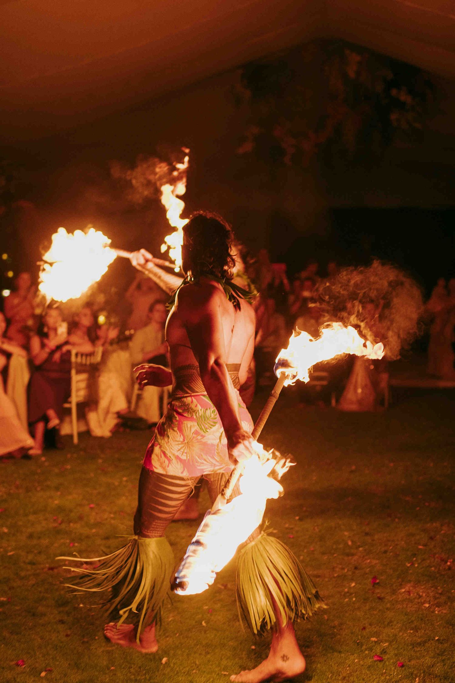 Fire dancer performing at night event, wearing grass skirt, twirling flaming torches at outdoor celebration.
