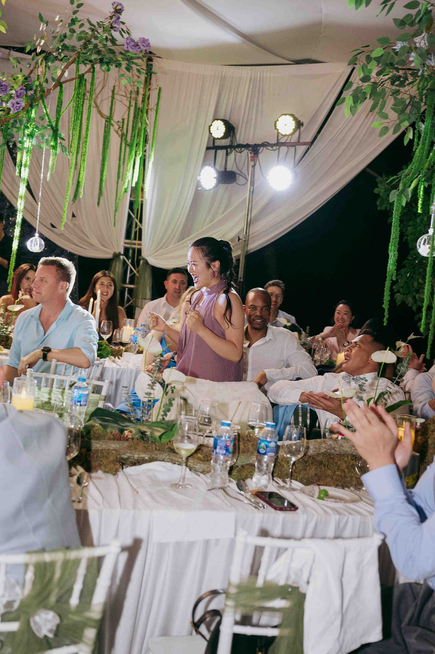 Guests laughing and celebrating at an elegant outdoor wedding reception with white drapes and green floral decor.