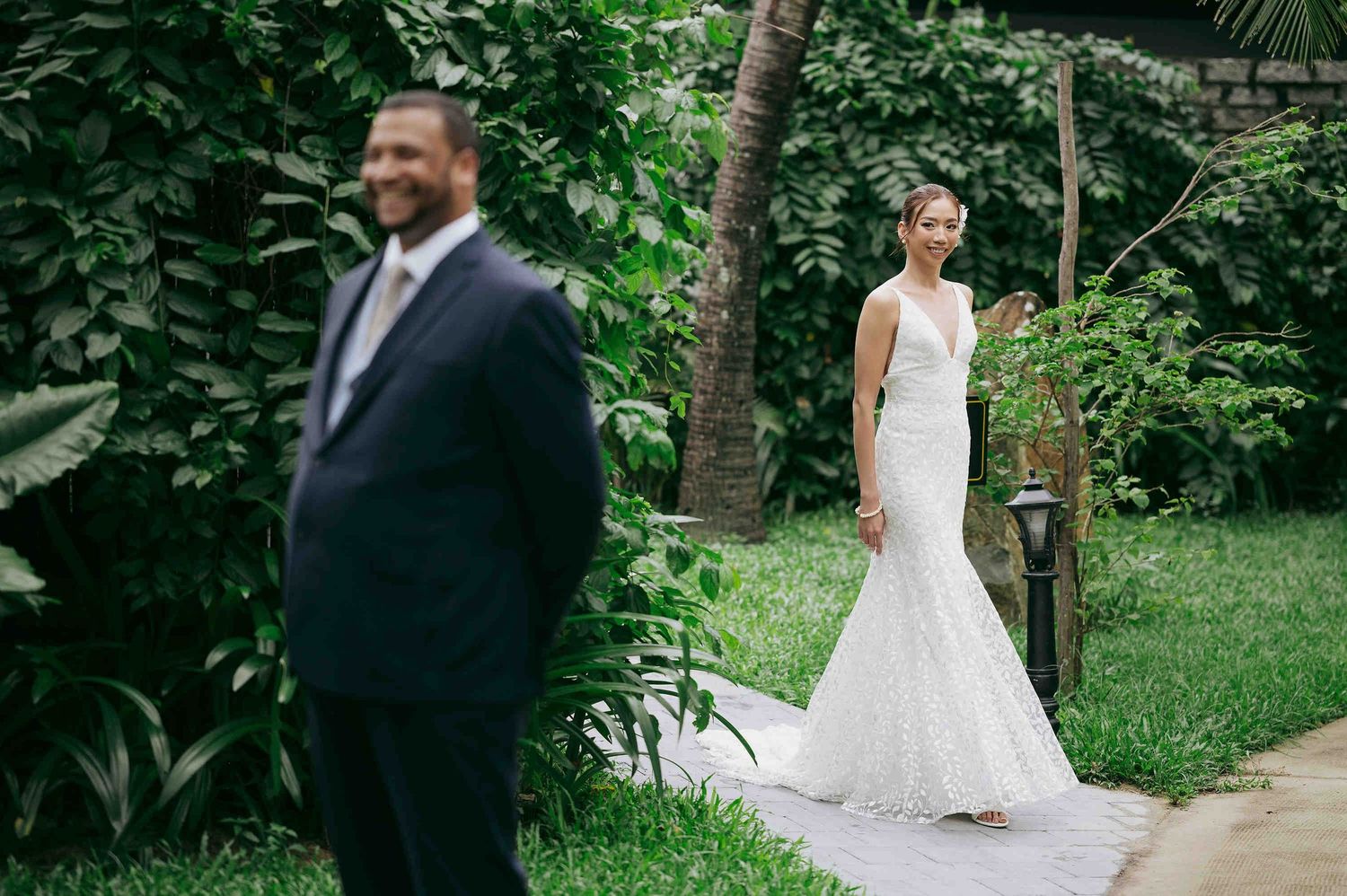 Bride in lace mermaid gown approaches groom in navy suit for first look in tropical garden setting.