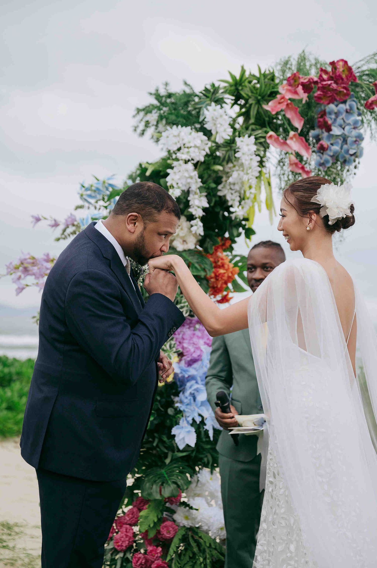 Groom kissing bride's hand at beach wedding ceremony under colorful floral arch with officiant.
