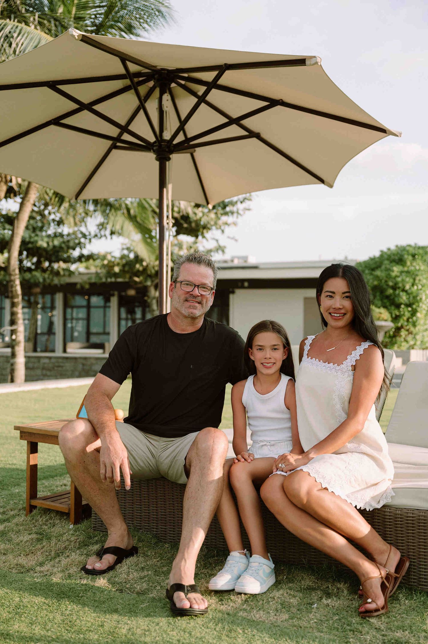 Da Nang family: three people sit together under a beige patio umbrella on outdoor furniture in a tropical garden setting, by AnhVuWedding.