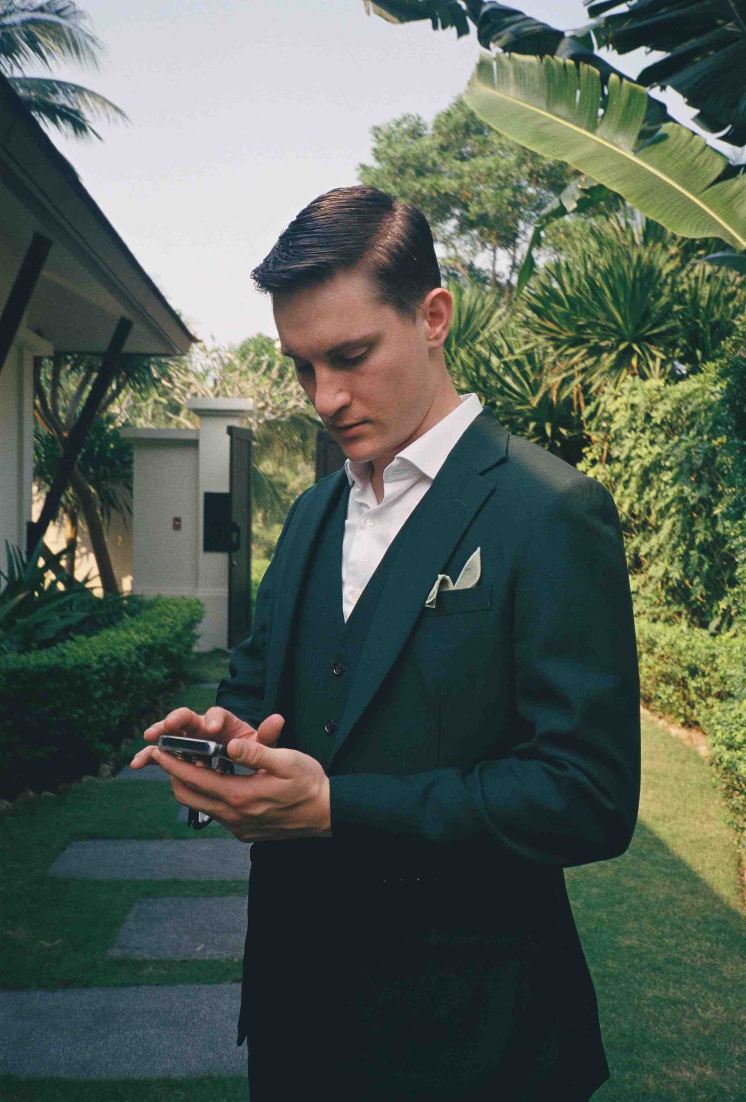 Lang Co wedding: person in a dark suit checks their phone while standing in a tropical garden setting, by AnhVuWedding.