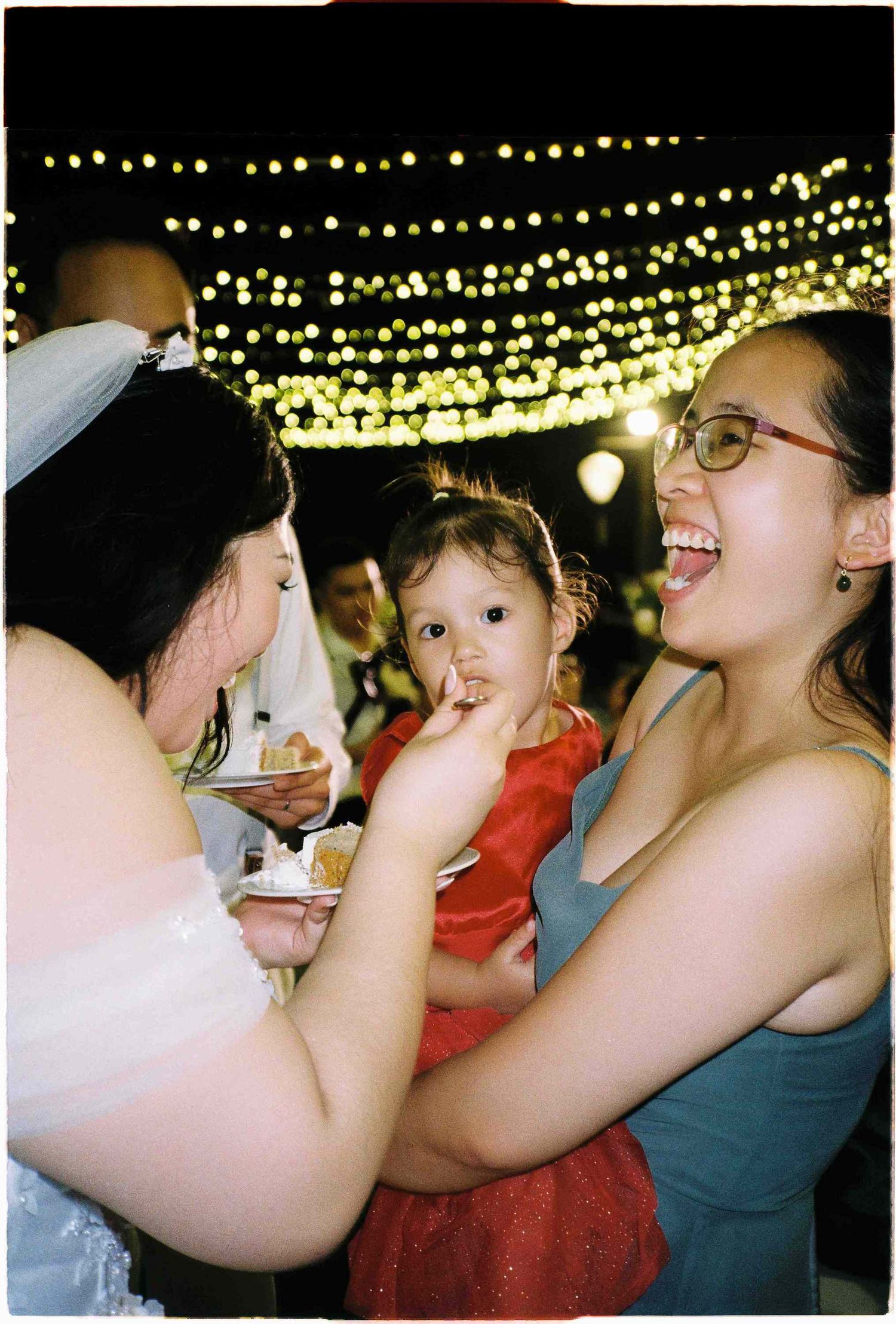 Bride feeding cake to a toddler in red dress held by smiling woman at outdoor wedding reception with fairy lights.