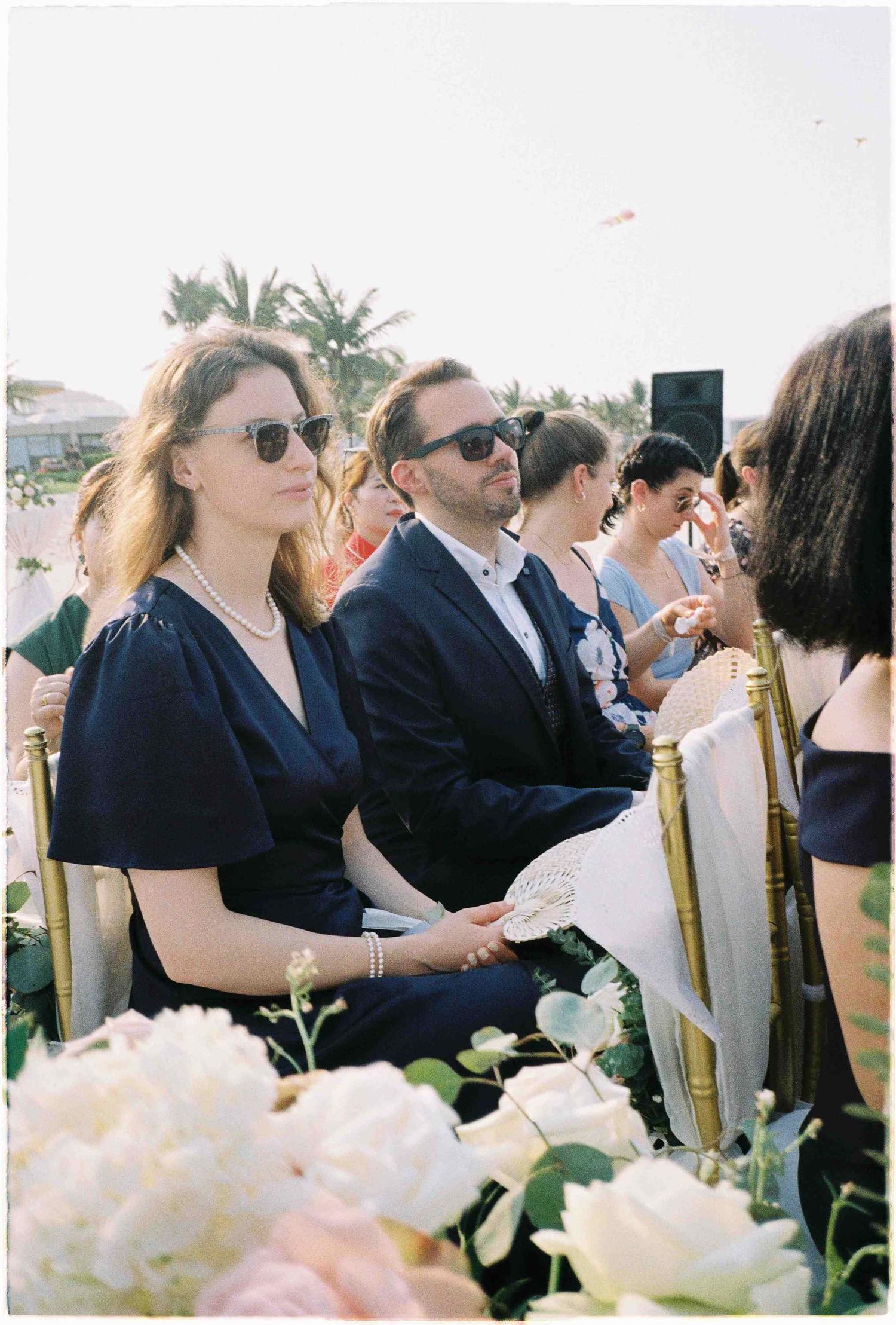 Guests in sunglasses seated at an outdoor wedding ceremony with white roses in the foreground.