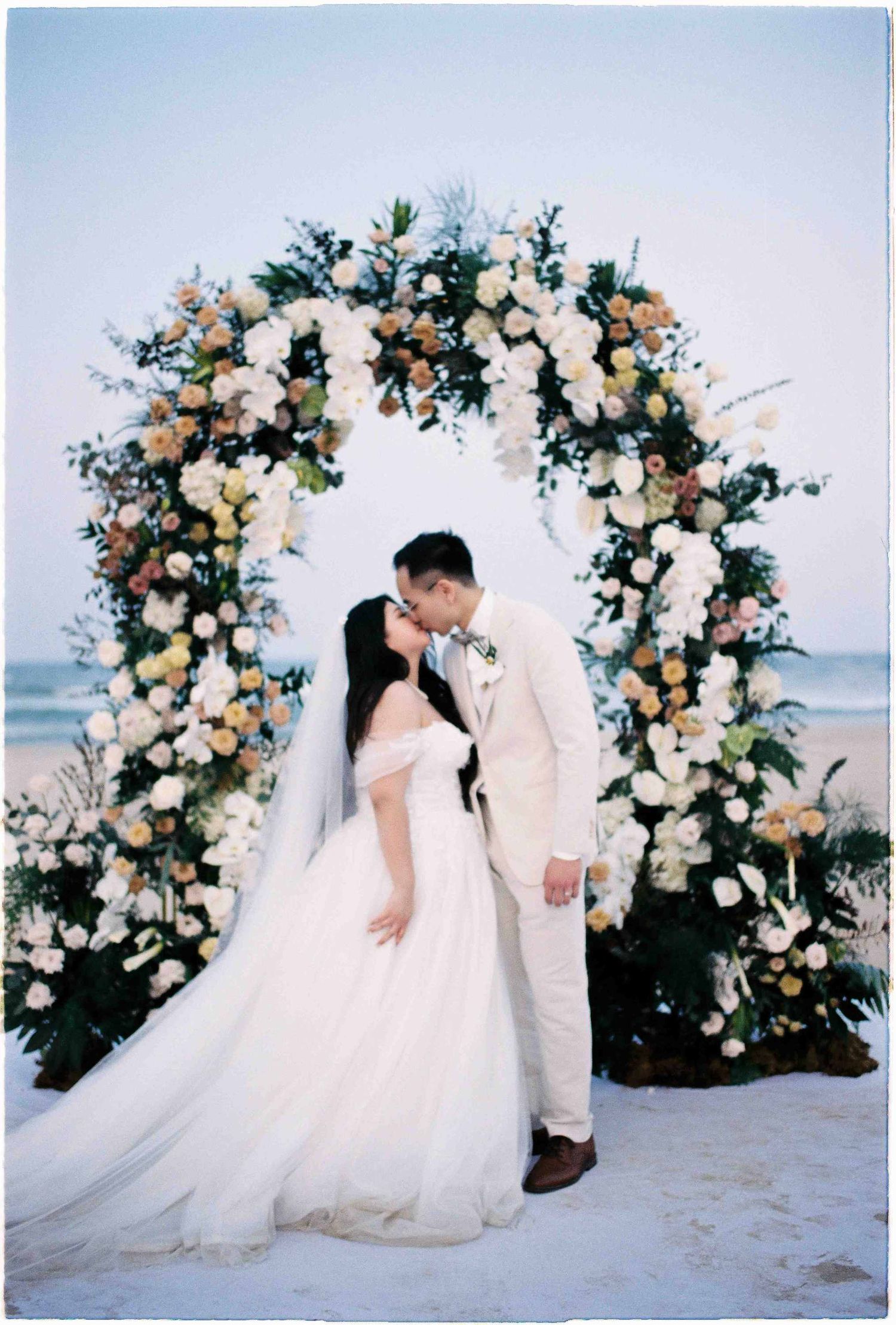 Couple kissing under a floral arch with white and yellow roses on a beach at a romantic outdoor wedding ceremony.
