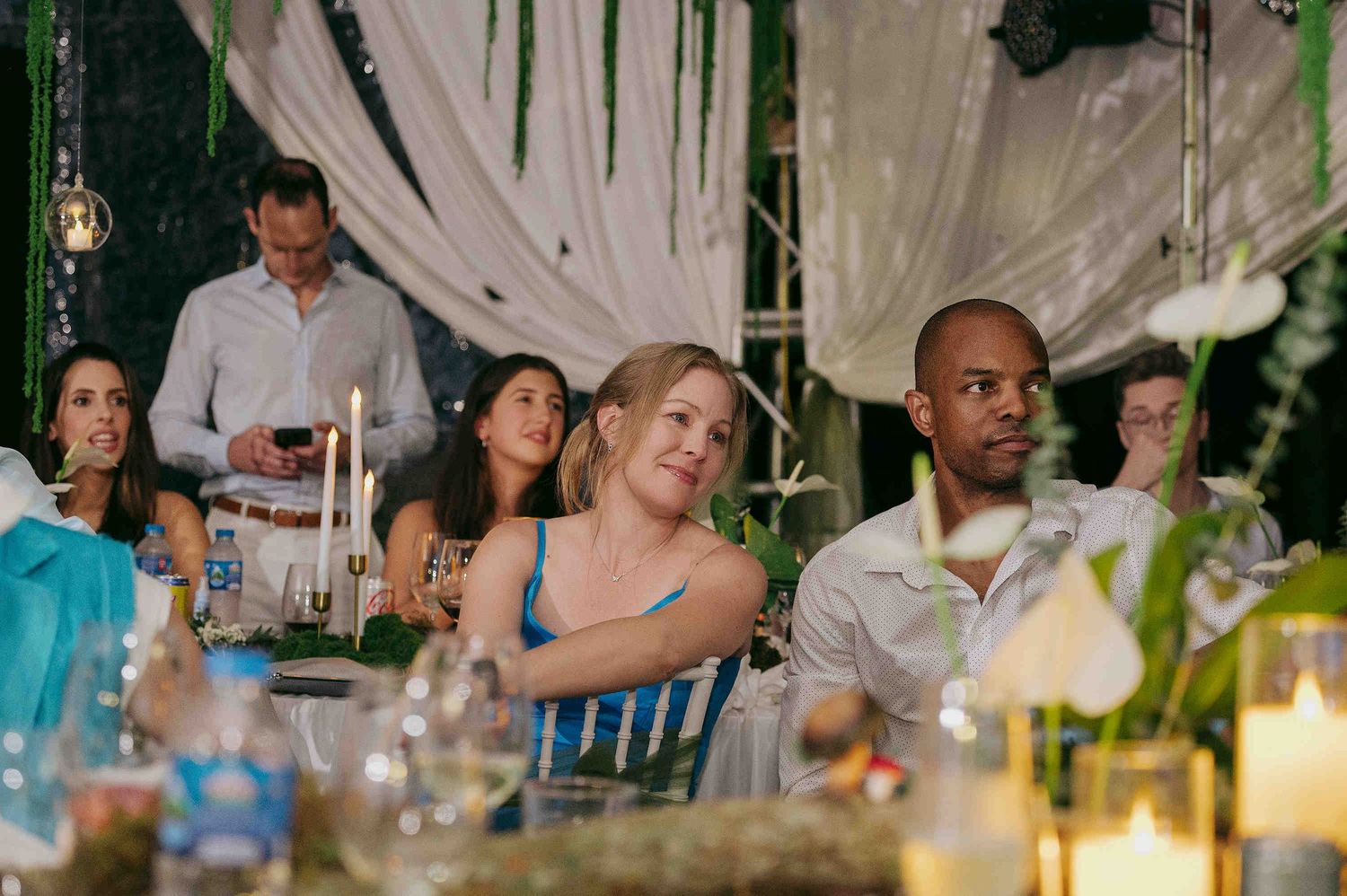 Guests seated at an elegant outdoor evening dinner party under draped canopy with candlelight and floral arrangements.