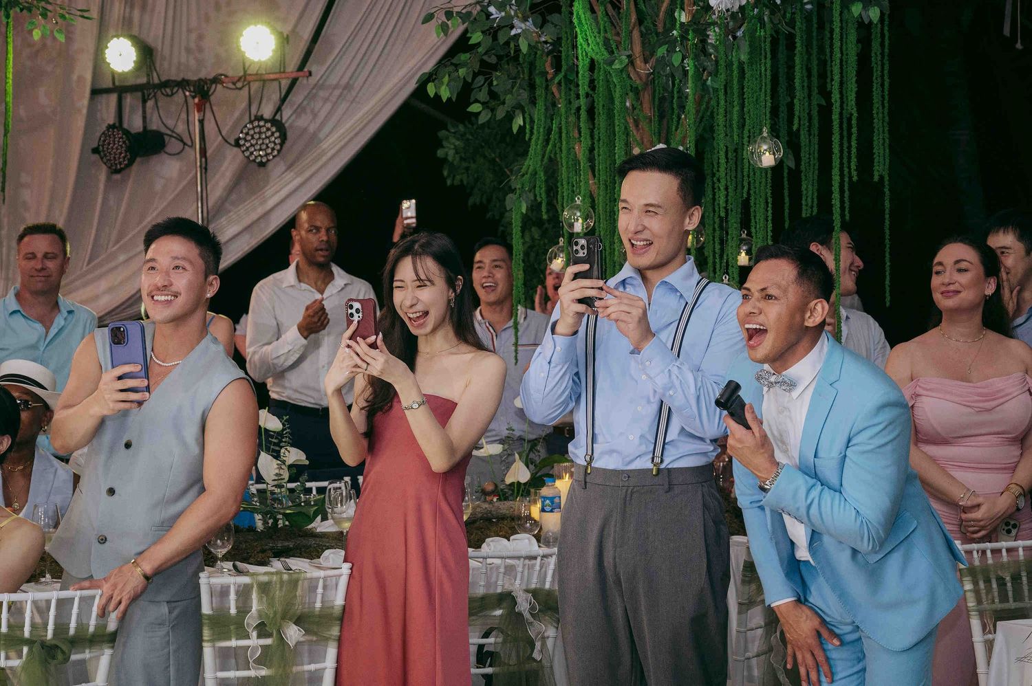 Guests laughing and taking photos at an outdoor wedding reception with lush green floral decorations.