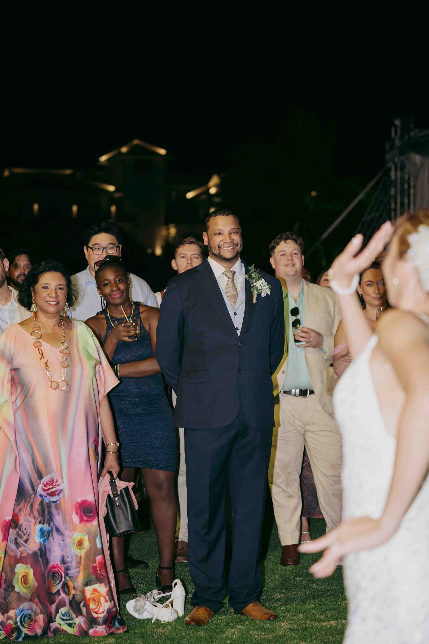 Guests gather outdoors at night watching a wedding bouquet toss, man in navy suit with boutonniere stands prominently.