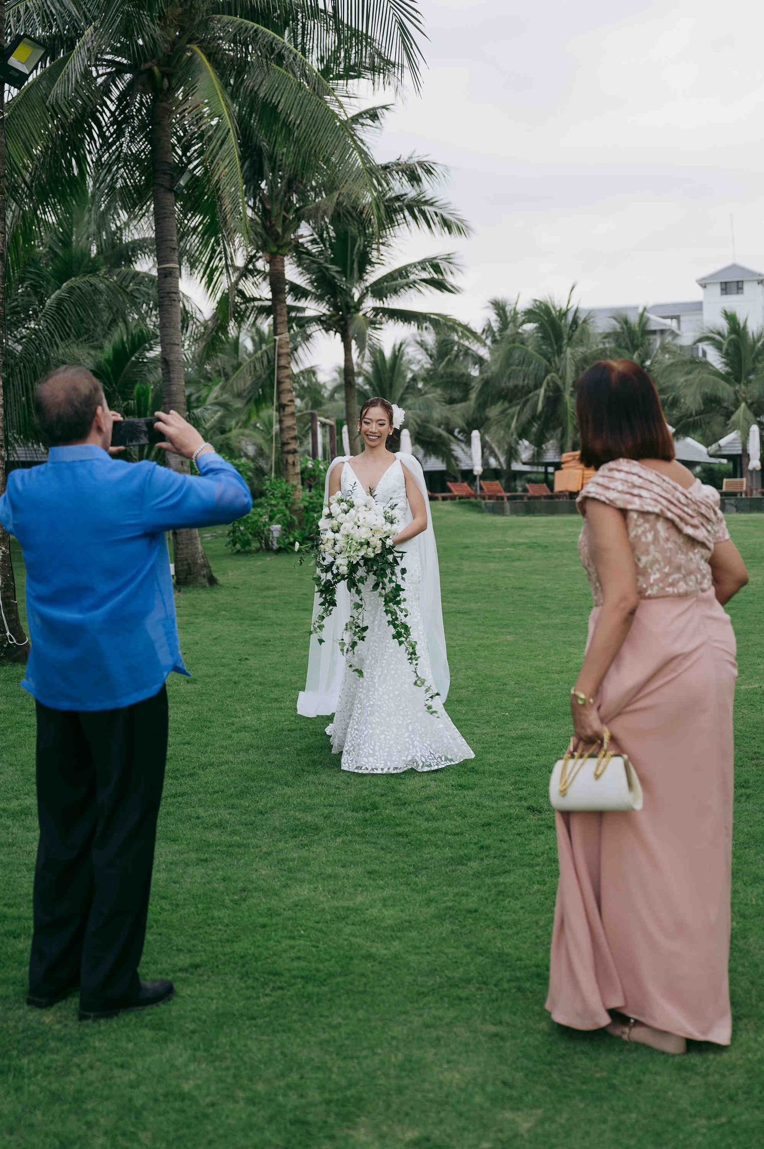 Bride in white gown holding bouquet poses on green lawn while guests photograph her at tropical beach wedding.