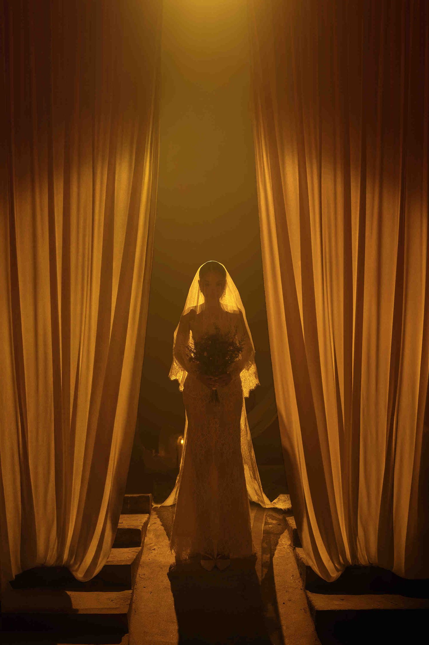 A bride in a white gown and veil stands between golden curtains, holding a bouquet in dramatic warm light.