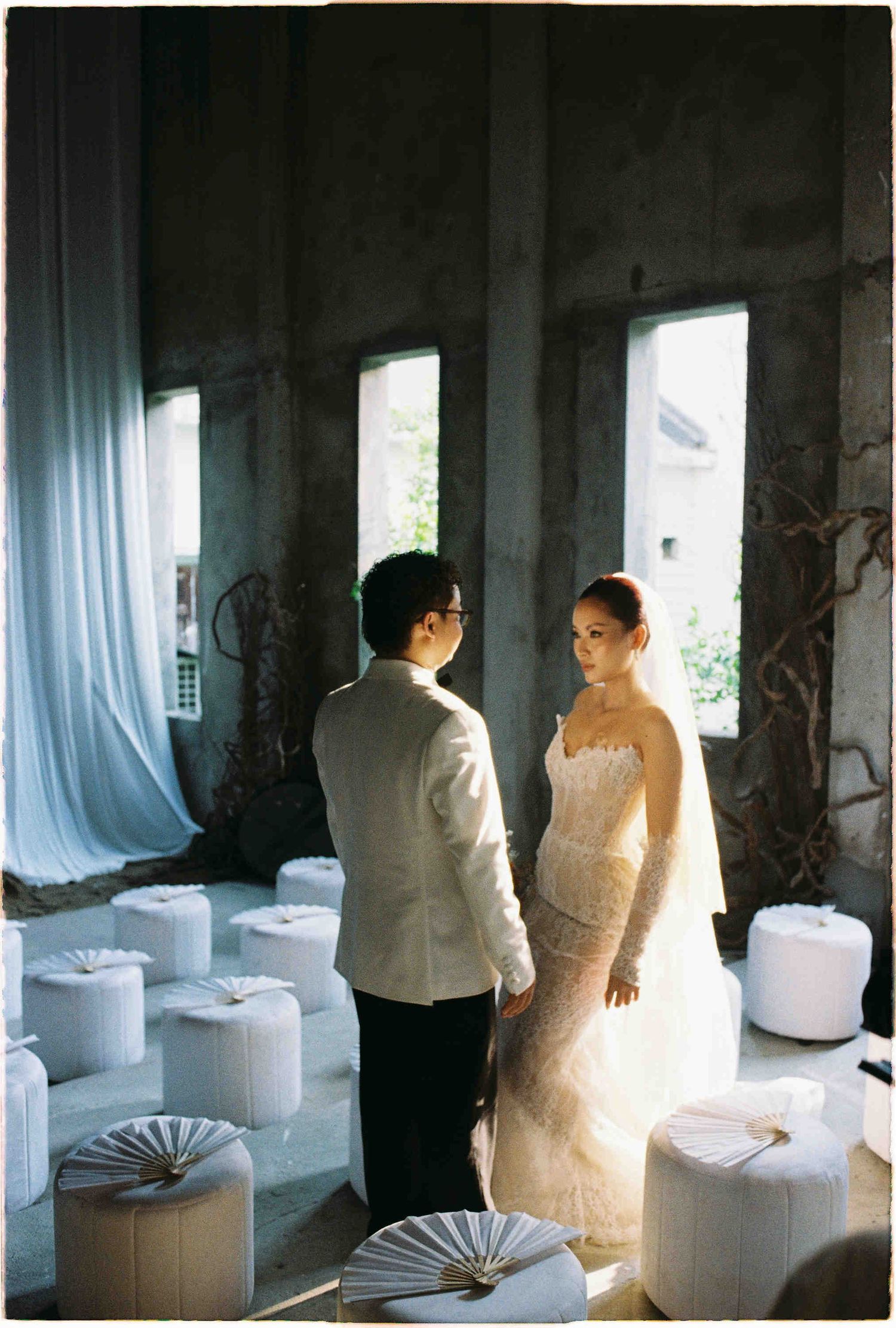 Bride in lace gown and groom in white suit stand facing each other in a sunlit rustic wedding venue with white decor.