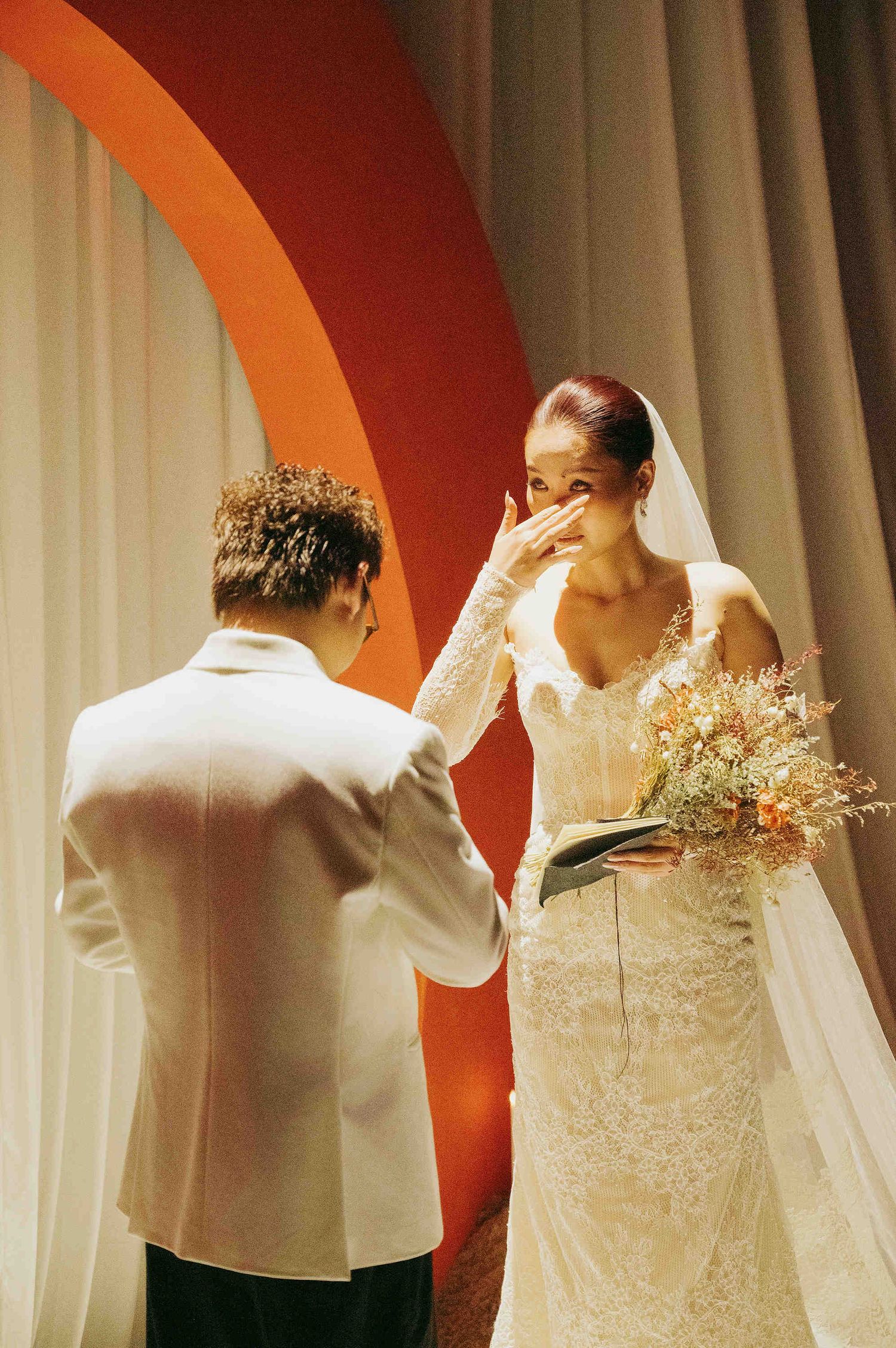 Emotional bride wiping tears in lace gown holding bouquet, facing groom in white suit at wedding ceremony.