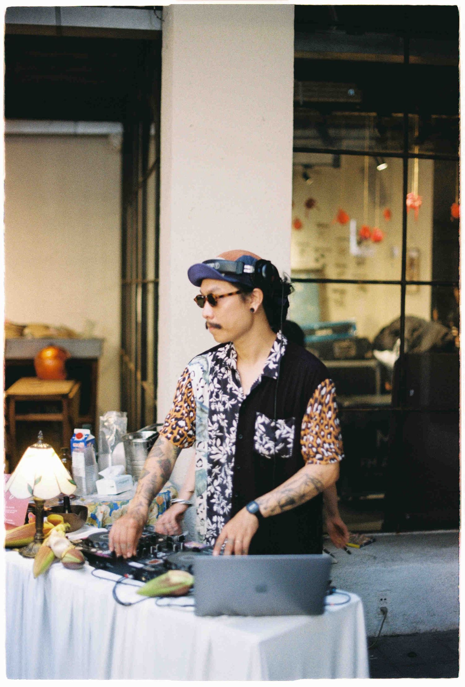 Tattooed DJ with cap and sunglasses mixing music outdoors on a laptop at a white table.