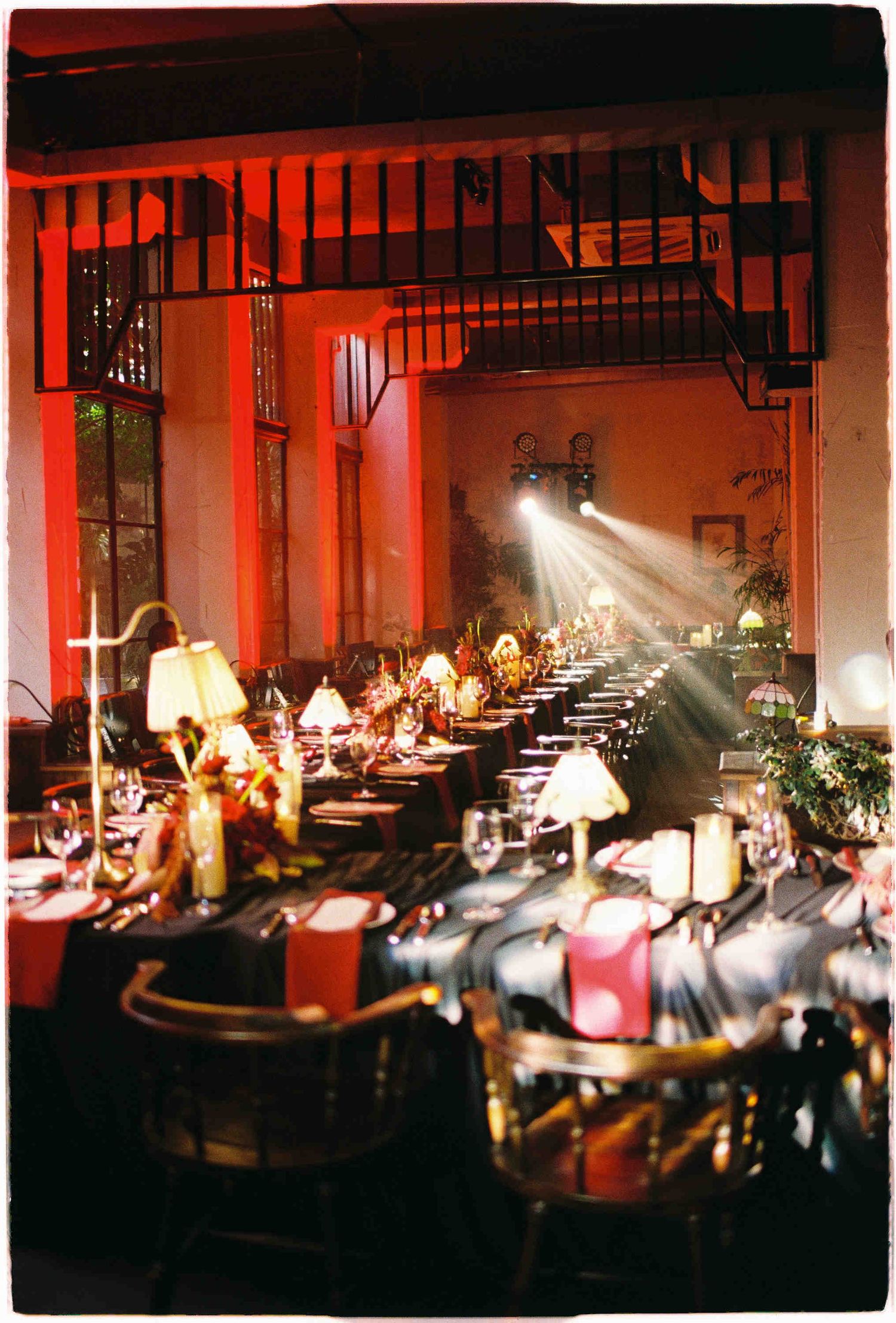 Elegant banquet hall with long dining tables, red ambient lighting, table lamps, and dramatic spotlight beams from above.