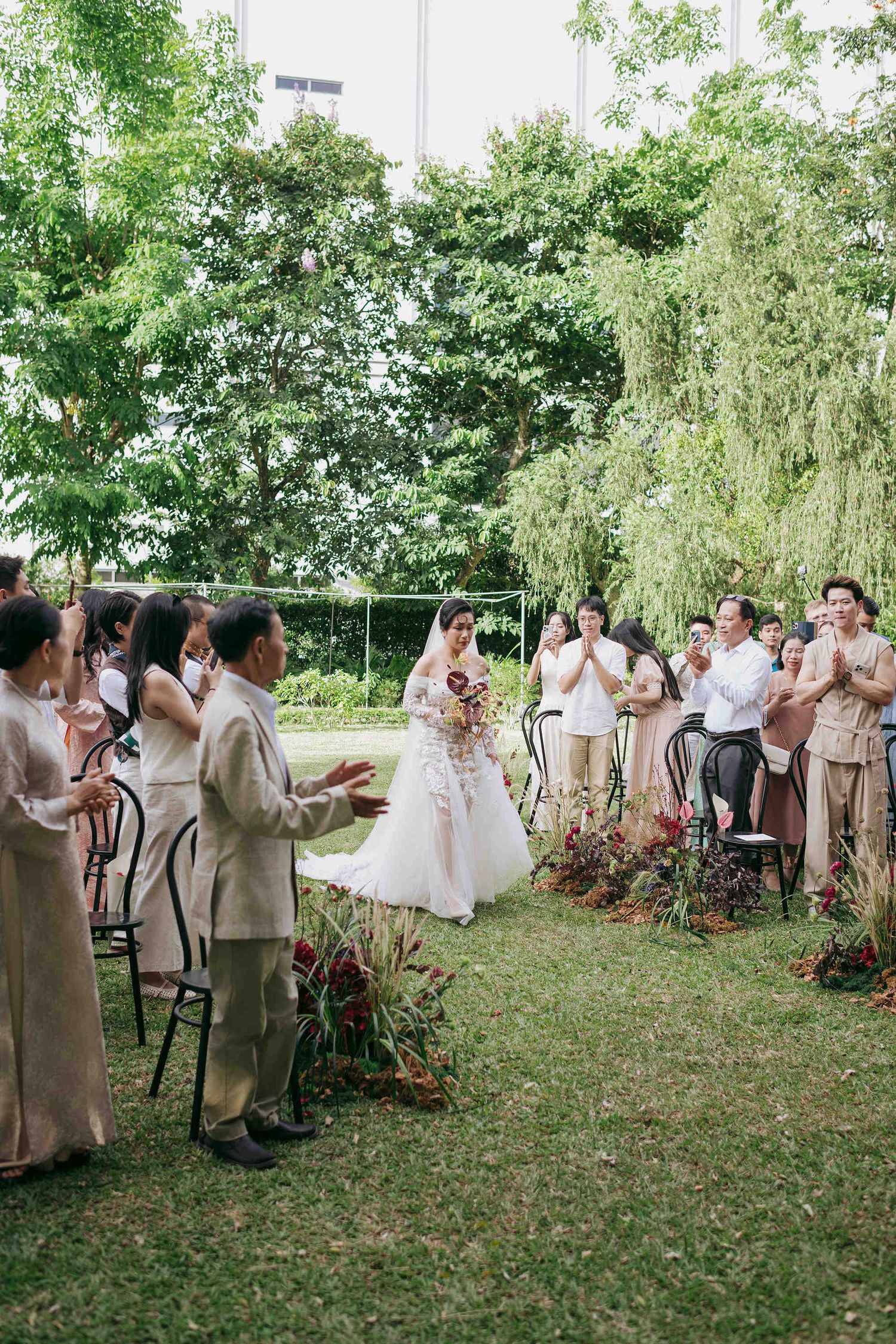 Bride walks down outdoor aisle in white gown as guests applaud at a garden wedding ceremony surrounded by lush green trees.