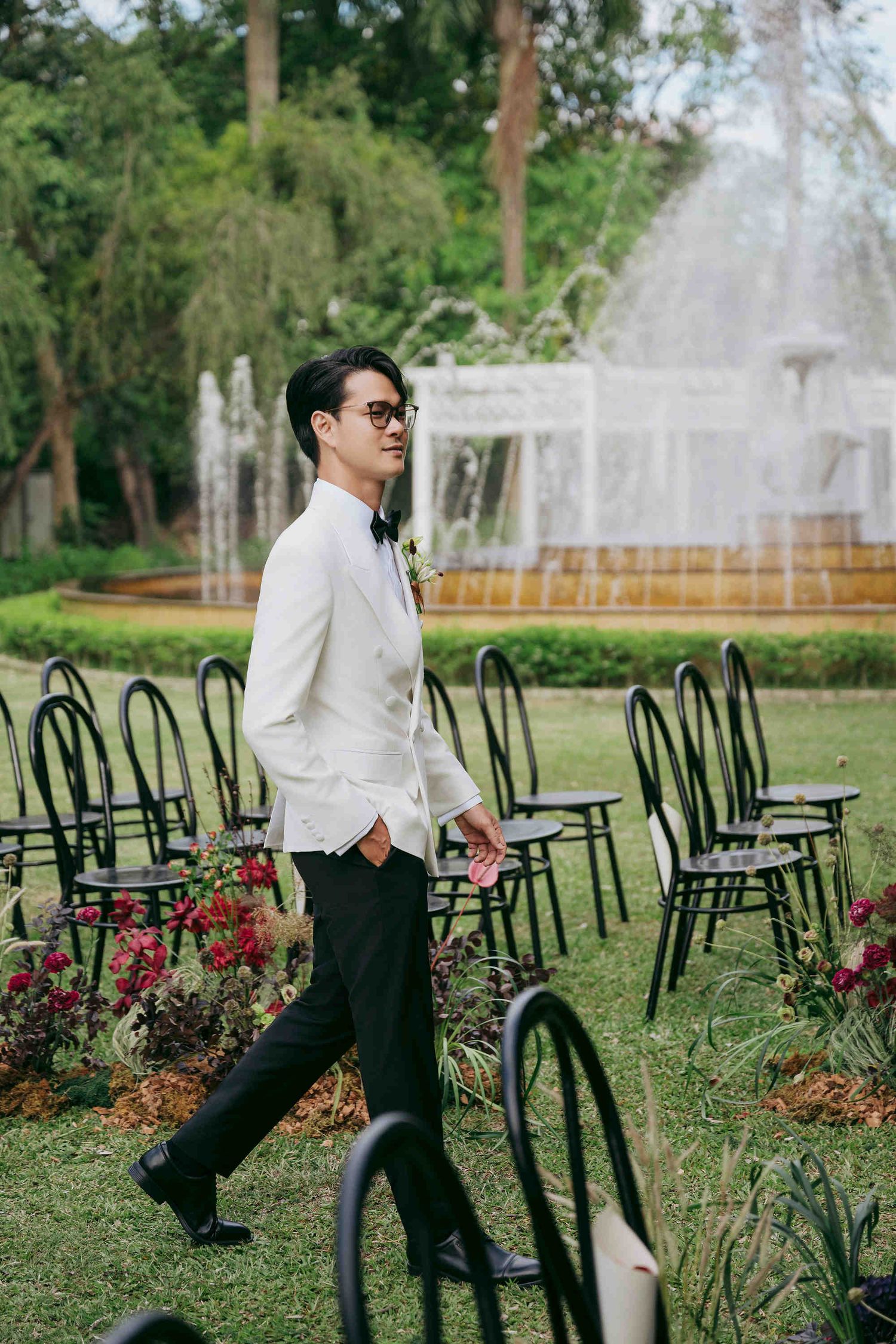 Groom in white jacket and sunglasses walks between black chairs at an outdoor garden wedding ceremony with fountain backdrop.
