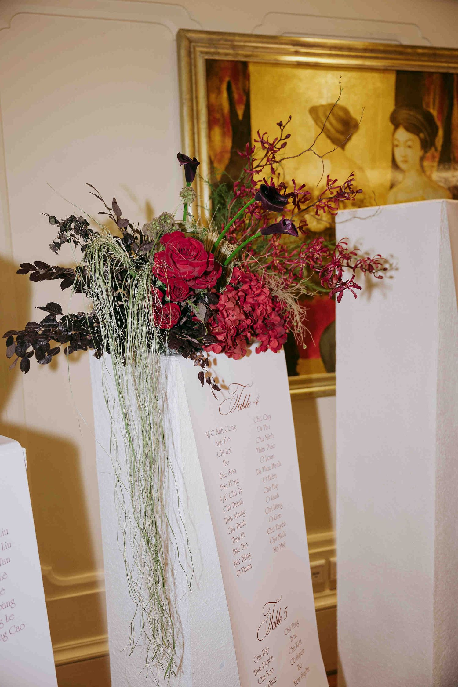 Elegant wedding seating chart display with lush red roses and hydrangea floral arrangement on white panels near a gold framed painting.