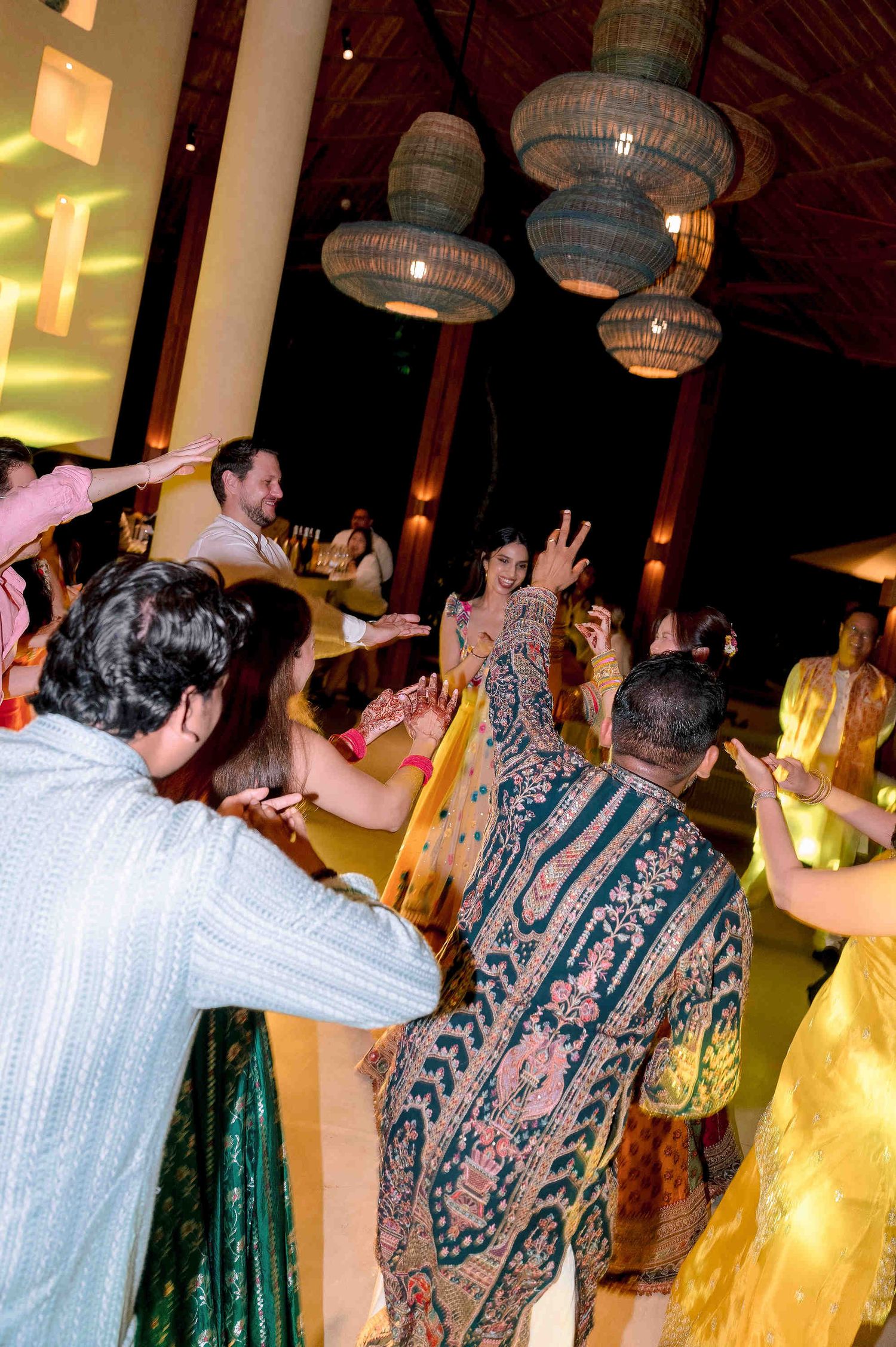 Guests in colorful traditional attire dance joyfully at an Indian wedding celebration under warm hanging lanterns.
