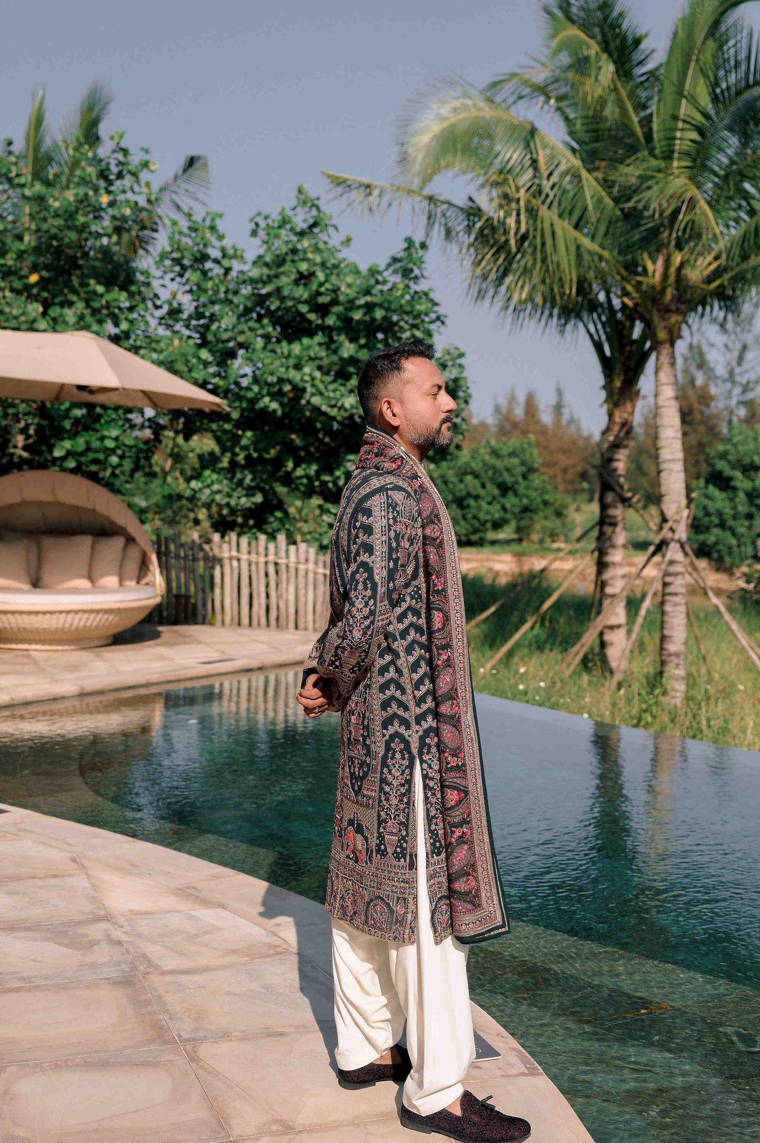Man in ornate embroidered sherwani stands by a pool surrounded by tropical greenery at an Indian wedding.