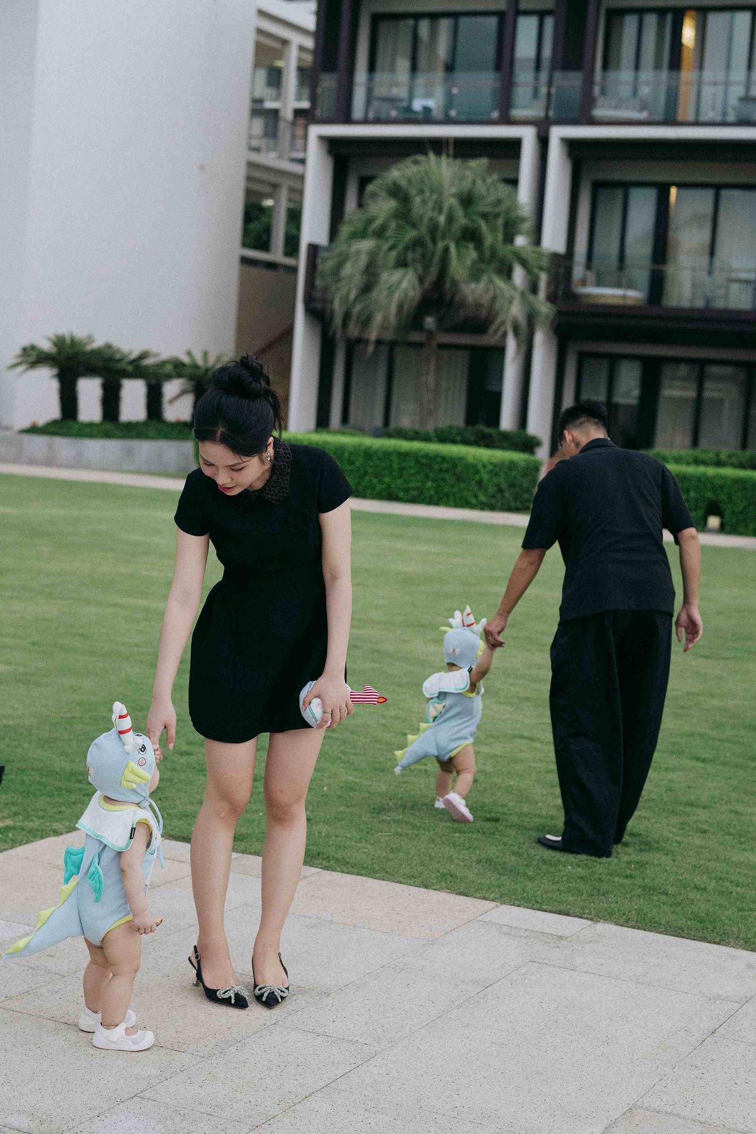 Da Nang family: toddler in a cute outfit follows adults along a paved walkway near modern buildings, by AnhVuWedding.