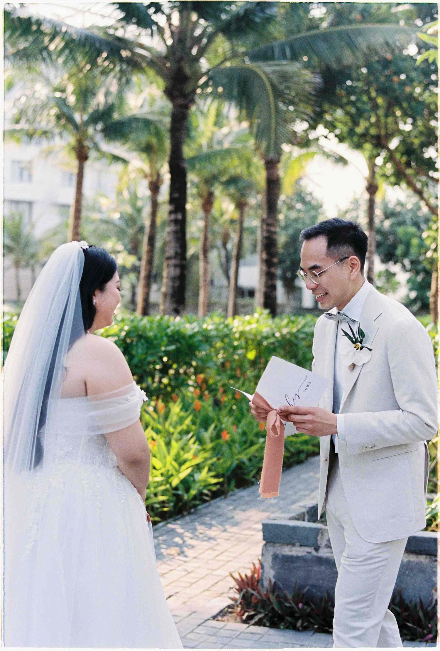 Groom reads wedding vows from a card to his bride in a white dress and veil, surrounded by tropical palm trees.