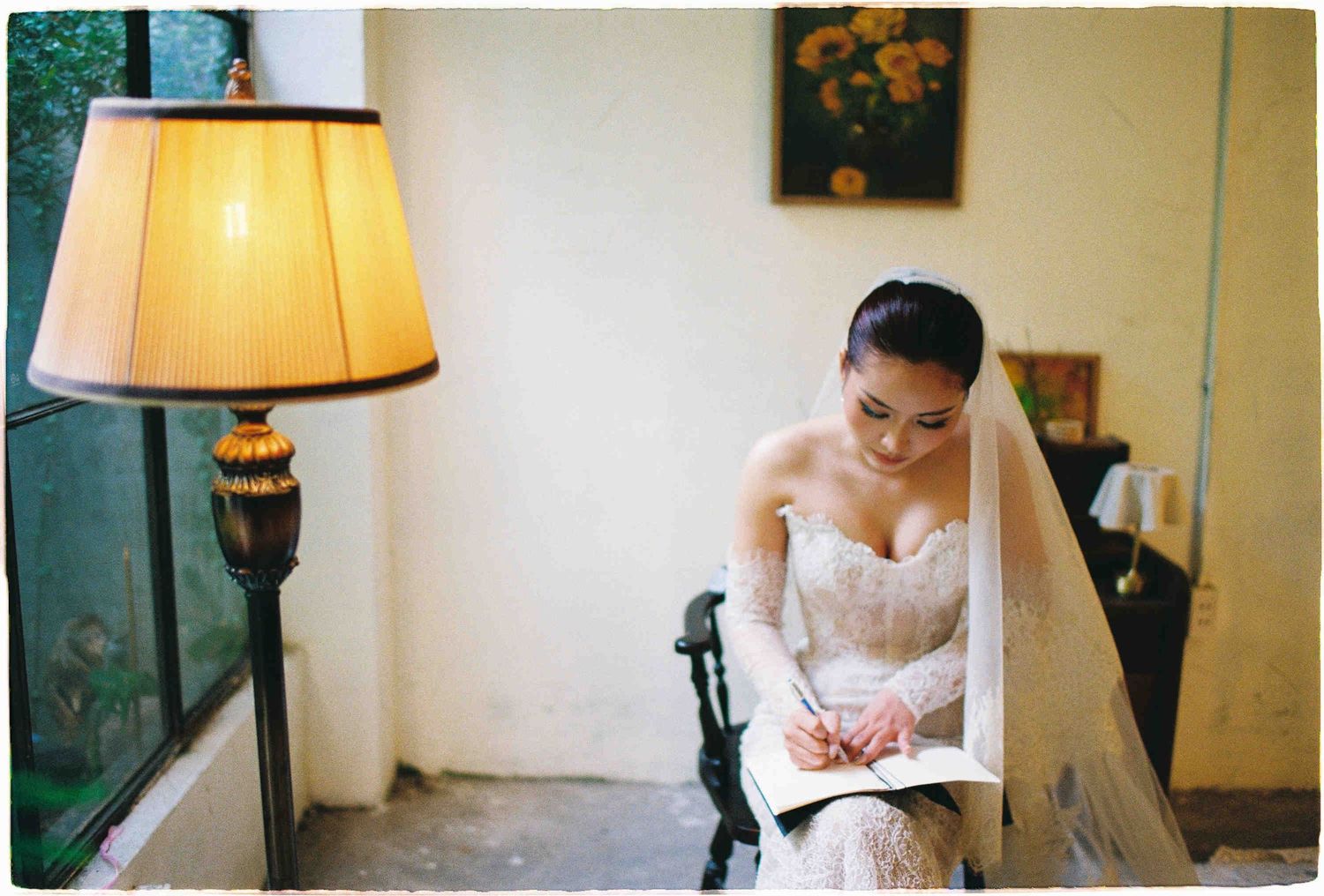 Bride in lace wedding dress and veil writing notes while seated near a vintage lamp in an elegant room.