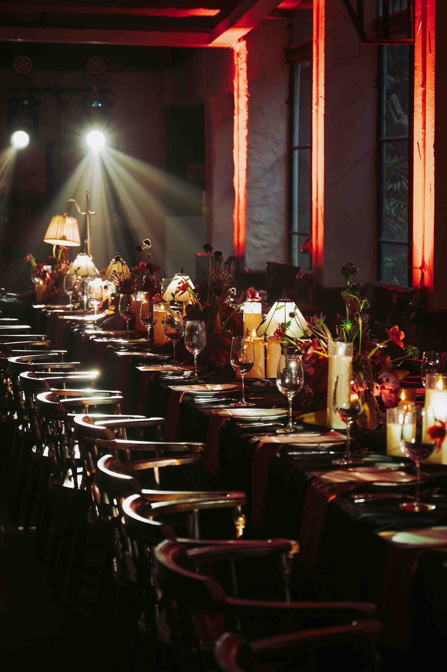 Elegant banquet hall with long dining tables, dark linens, candlelight, floral centerpieces, and dramatic red uplighting.