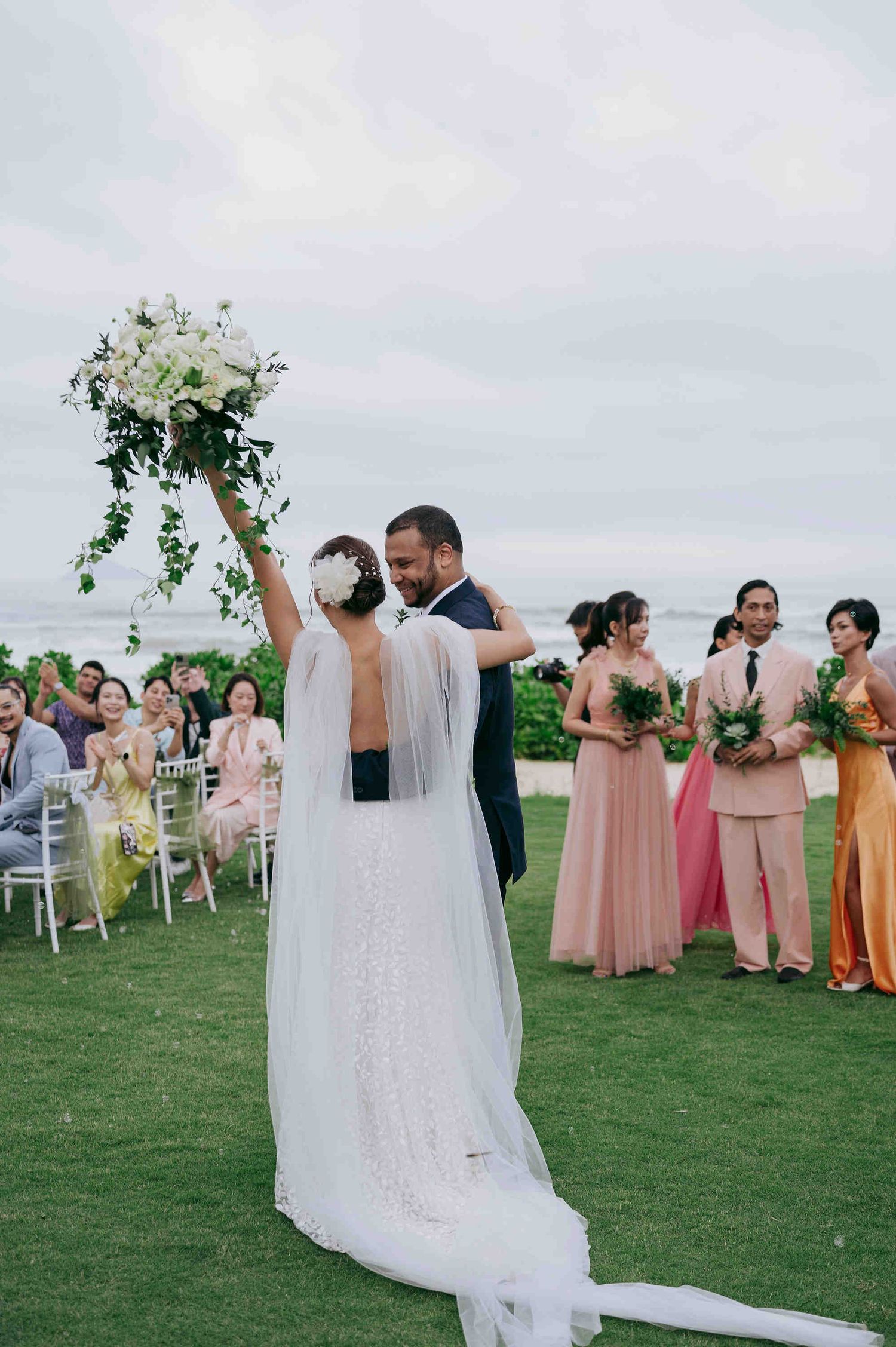 Bride raises bouquet of white flowers as groom embraces her, bridesmaids in pink dresses stand nearby at outdoor wedding.