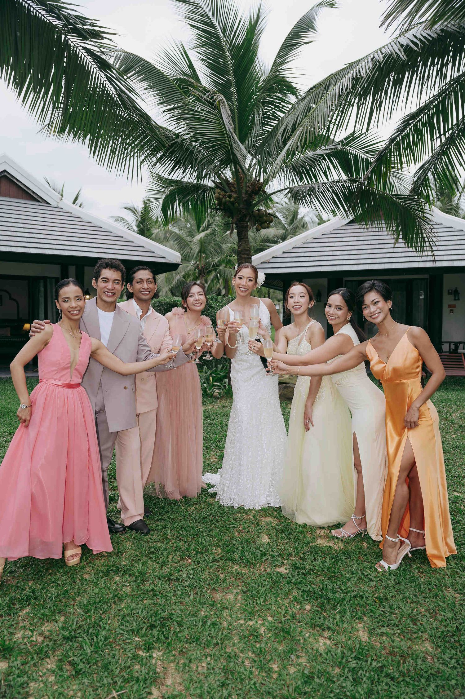 Bride in white gown poses with bridesmaids in colorful dresses and guests, toasting outdoors near palm trees.