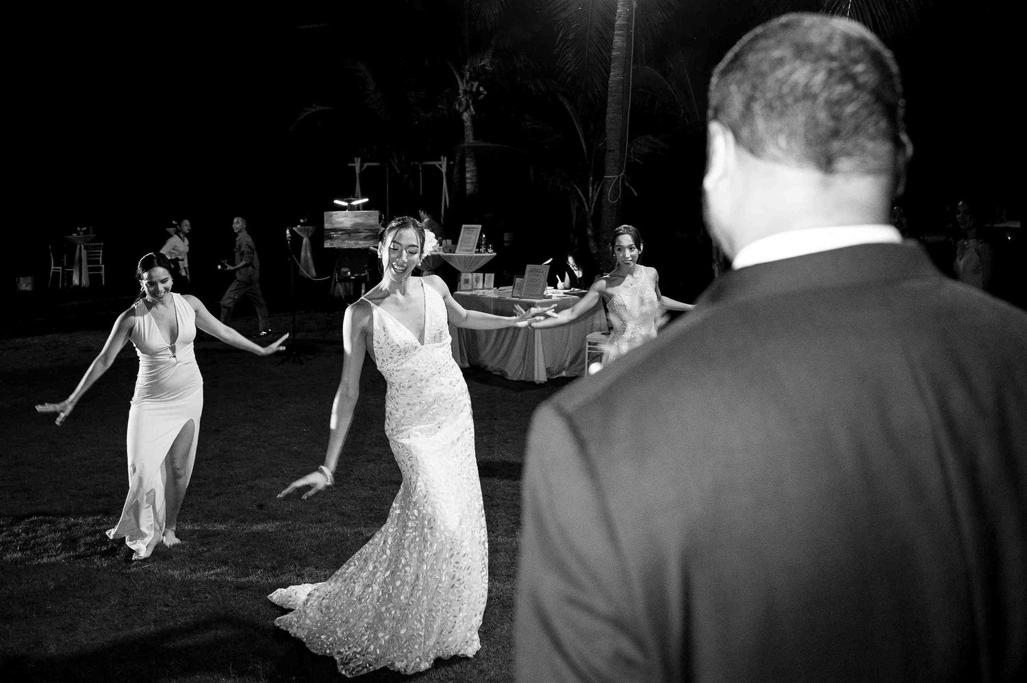 Bride in lace gown dancing joyfully with guests at an outdoor evening wedding reception in black and white photo.