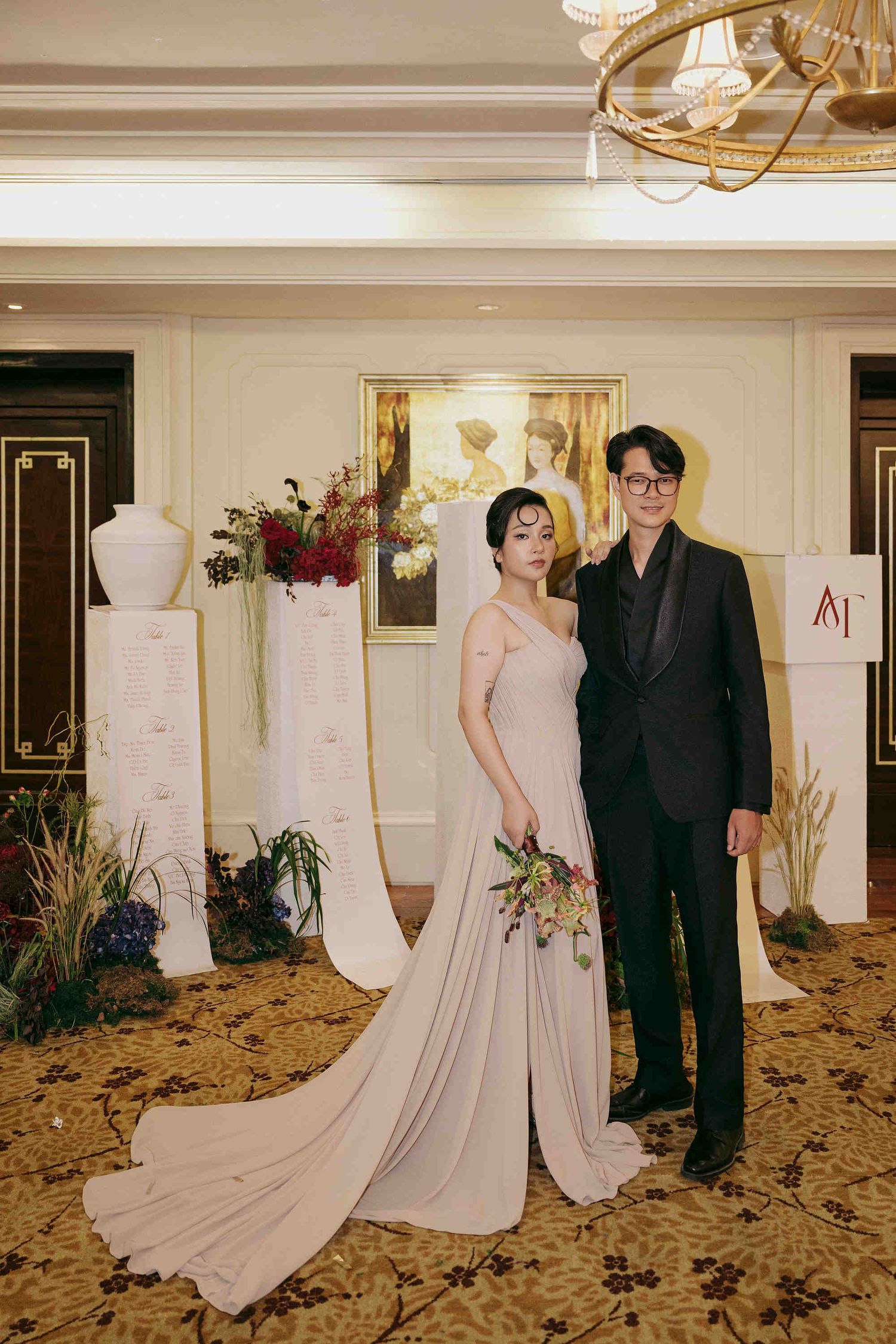 Couple posing at elegant wedding venue, woman in blush gown holding bouquet, man in black suit.