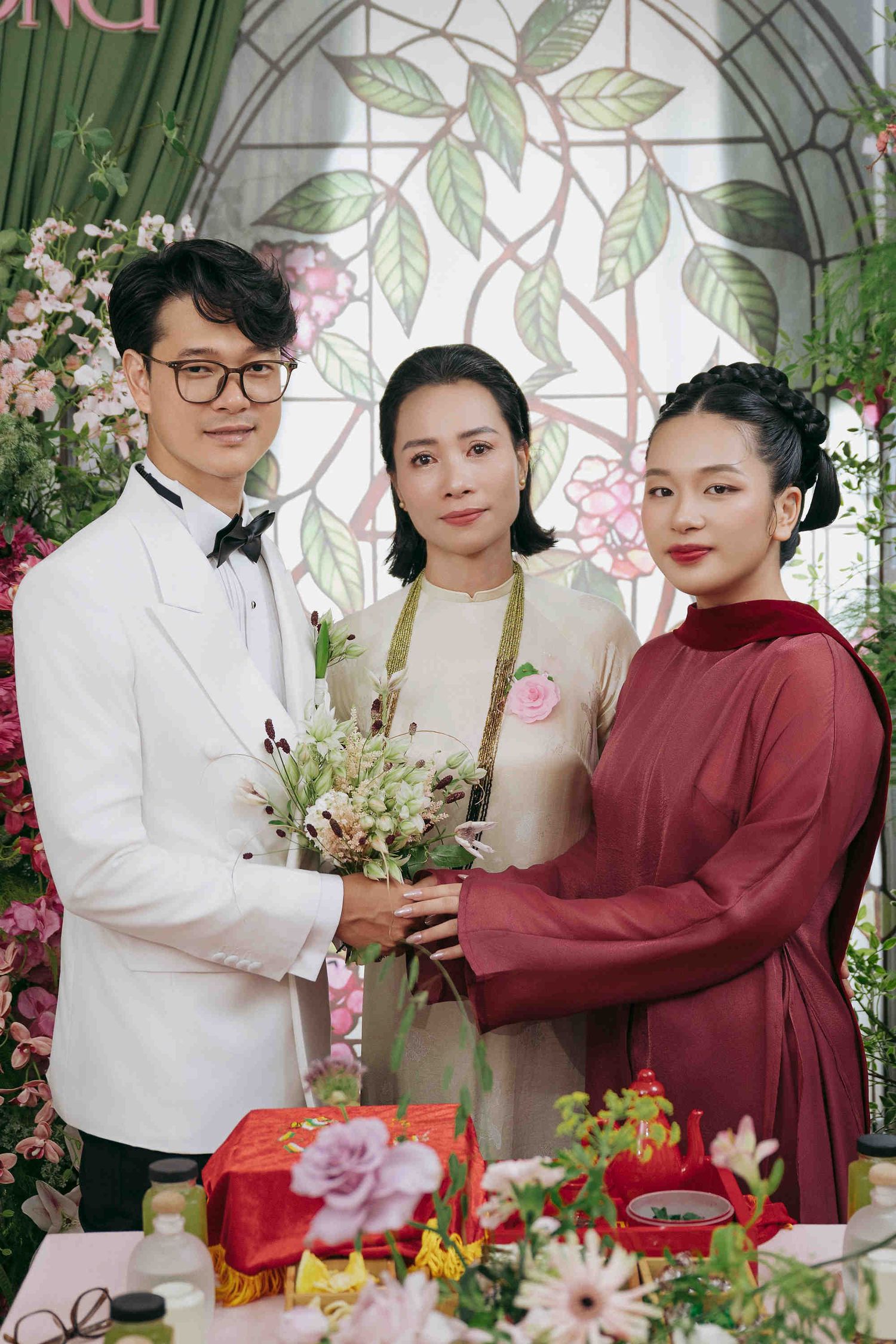 Three people pose at a traditional Vietnamese wedding ceremony with floral decorations and red gift boxes.
