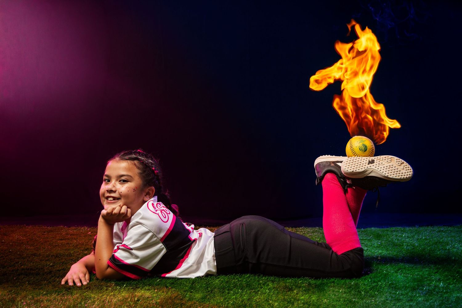 Young girl in softball uniform lies on grass smiling, with a softball balanced on a shoe emitting dramatic flames.