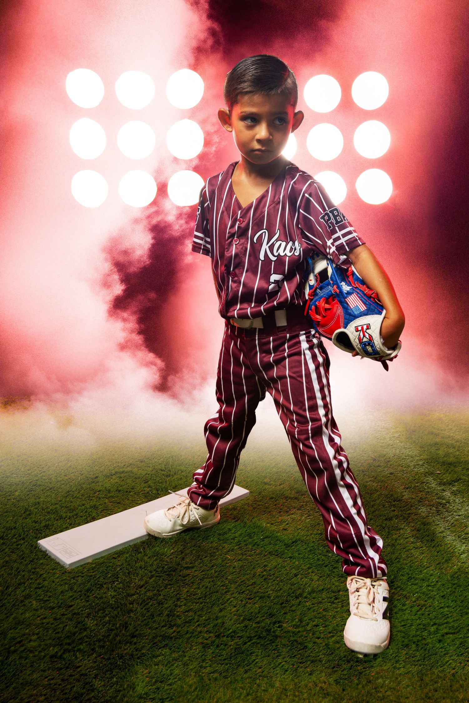 Young boy in maroon pinstripe baseball uniform holding glove, standing on pitchers mound with dramatic stadium lights and red smoke.
