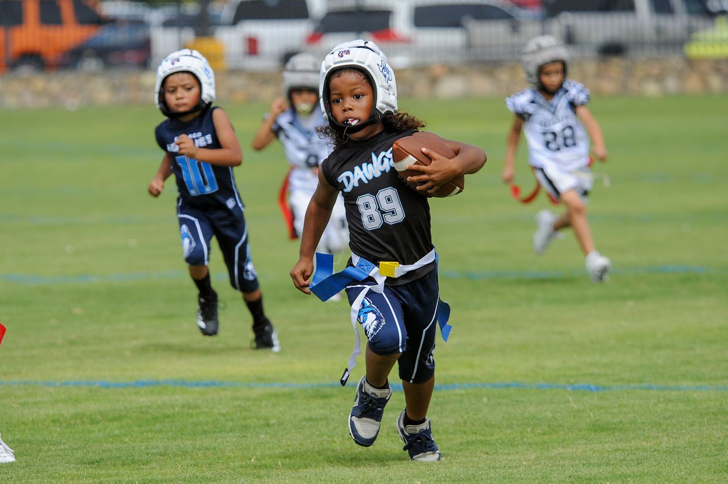 Young football players in black uniforms racing on a green field during a youth league game, number 89 carrying the ball.