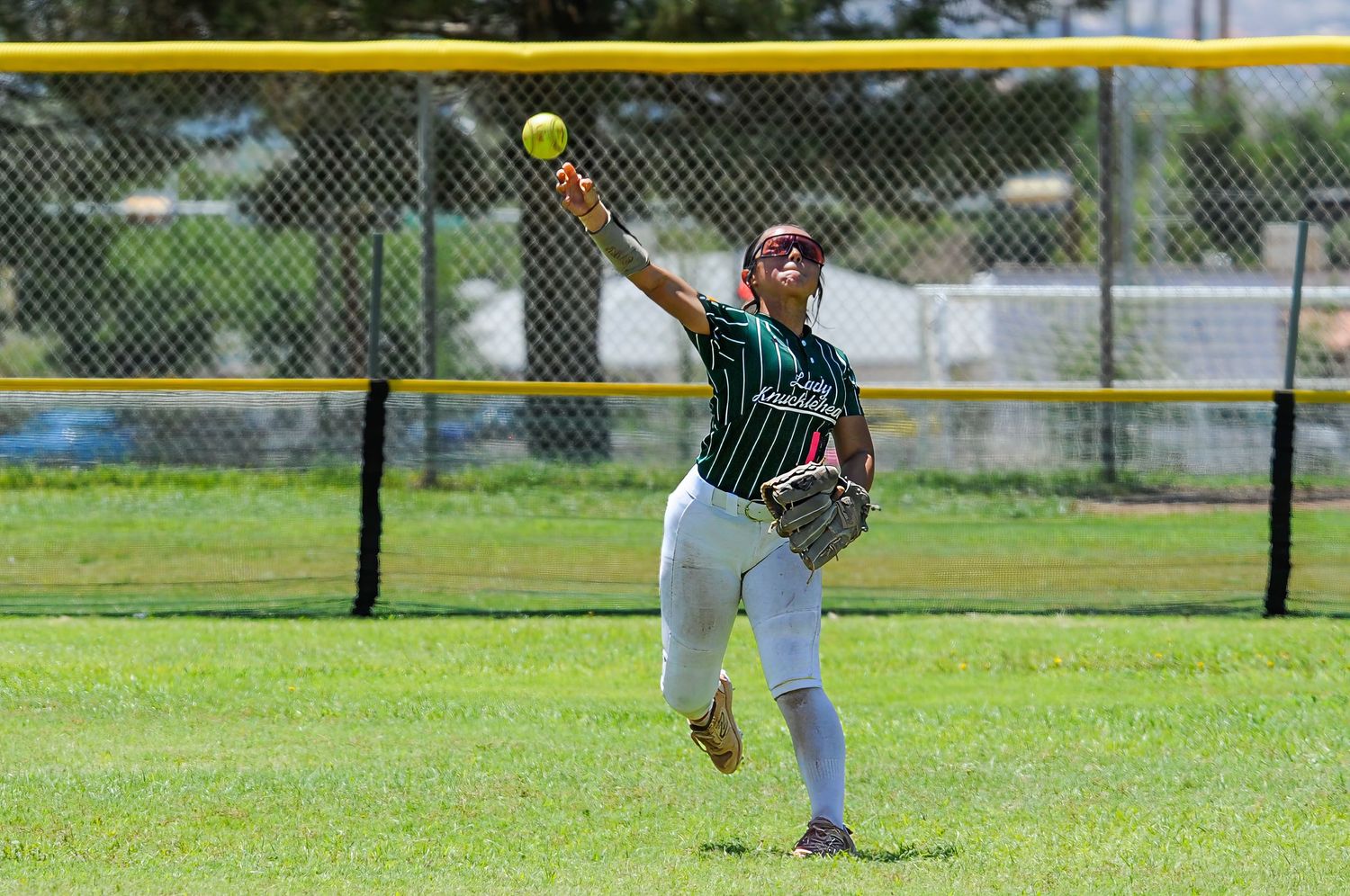 Female softball outfielder in green uniform catching a fly ball near the outfield fence on a sunny day.