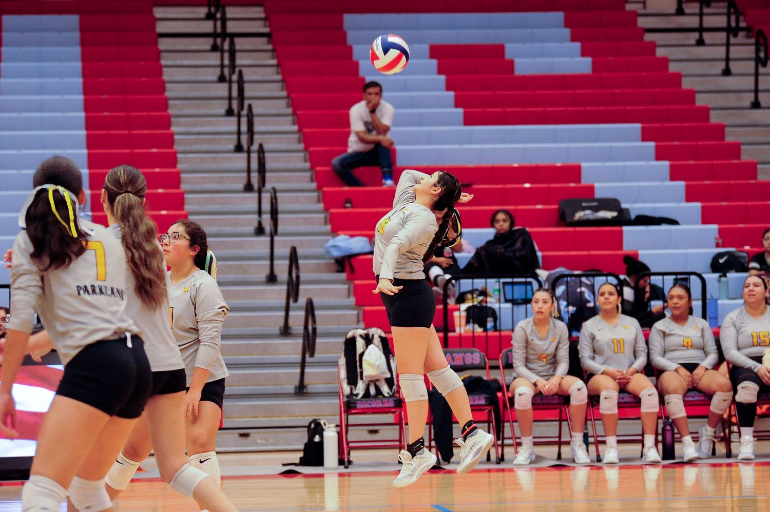 Female volleyball player leaps to spike the ball during an indoor match with teammates watching nearby.