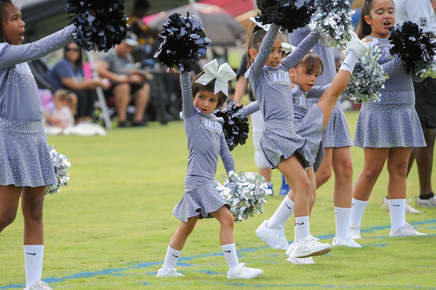 Young cheerleaders in gray uniforms perform with silver pom-poms on a green field during a youth cheerleading event.