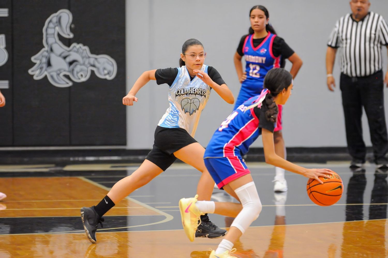 Young girls playing competitive basketball game, defender guarding dribbler on indoor court.