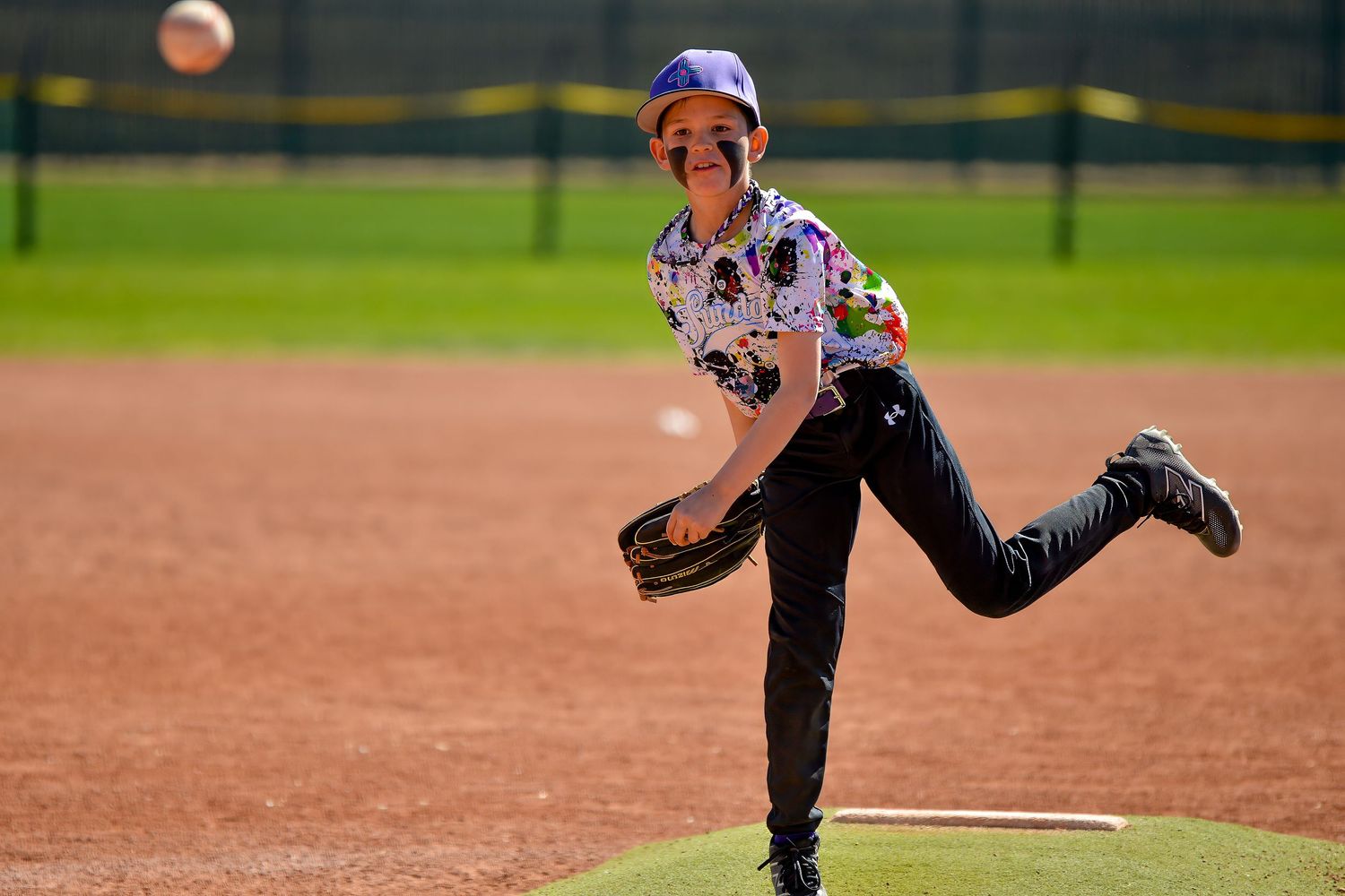 Smiling young baseball pitcher in patterned uniform delivers a pitch from the mound.