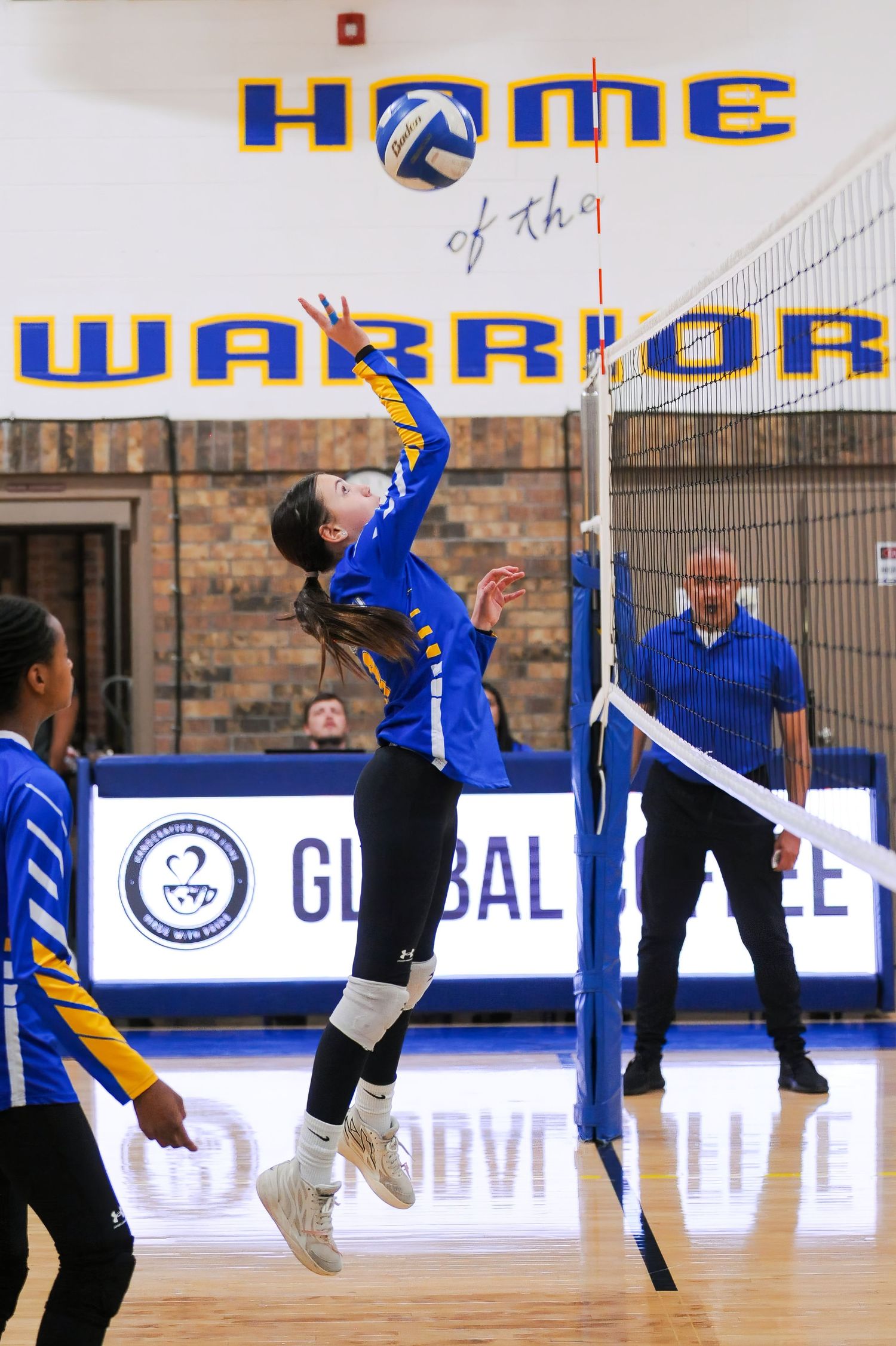 A female volleyball player in blue uniform jumps to spike the ball over the net in a Warriors gymnasium.