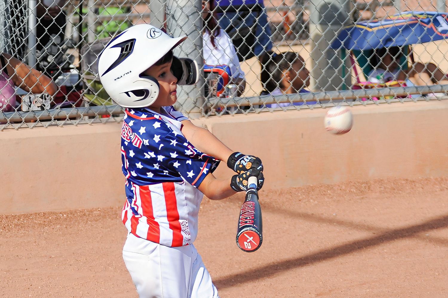 Young baseball player in patriotic stars and stripes uniform making contact with pitch during game.