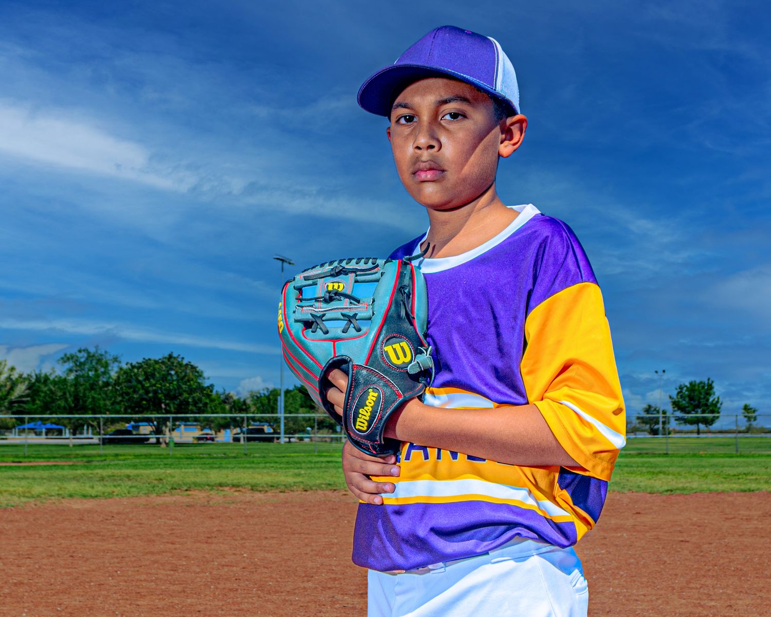 Young boy in purple baseball uniform holding a Wilson glove on a baseball field under a blue sky.