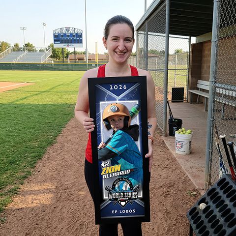Woman smiling holding a framed baseball World Series banner for Zion Alvarado, EP Lobos team, at a baseball field.