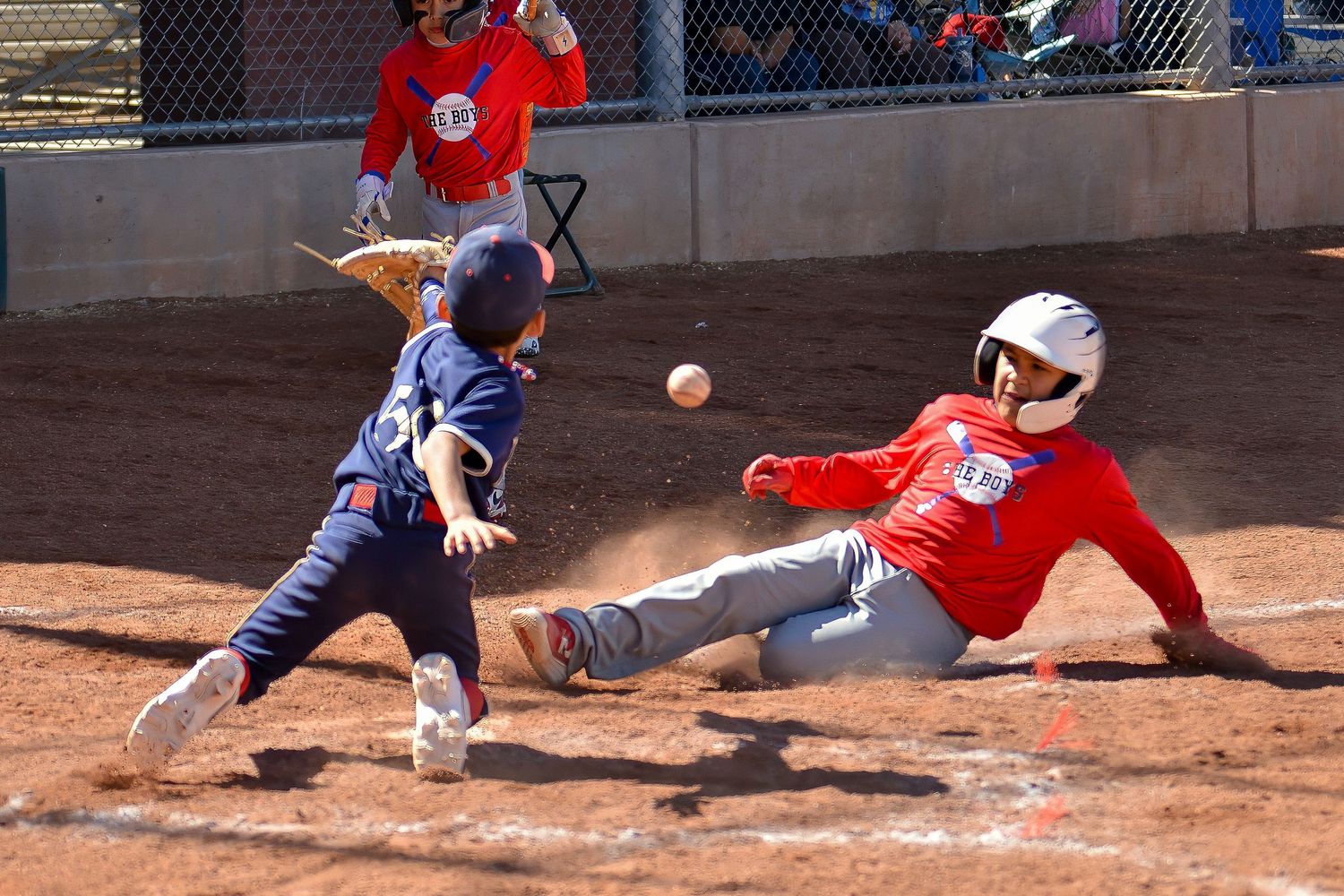 Little league runner slides into home plate as catcher reaches for the ball in a close play.