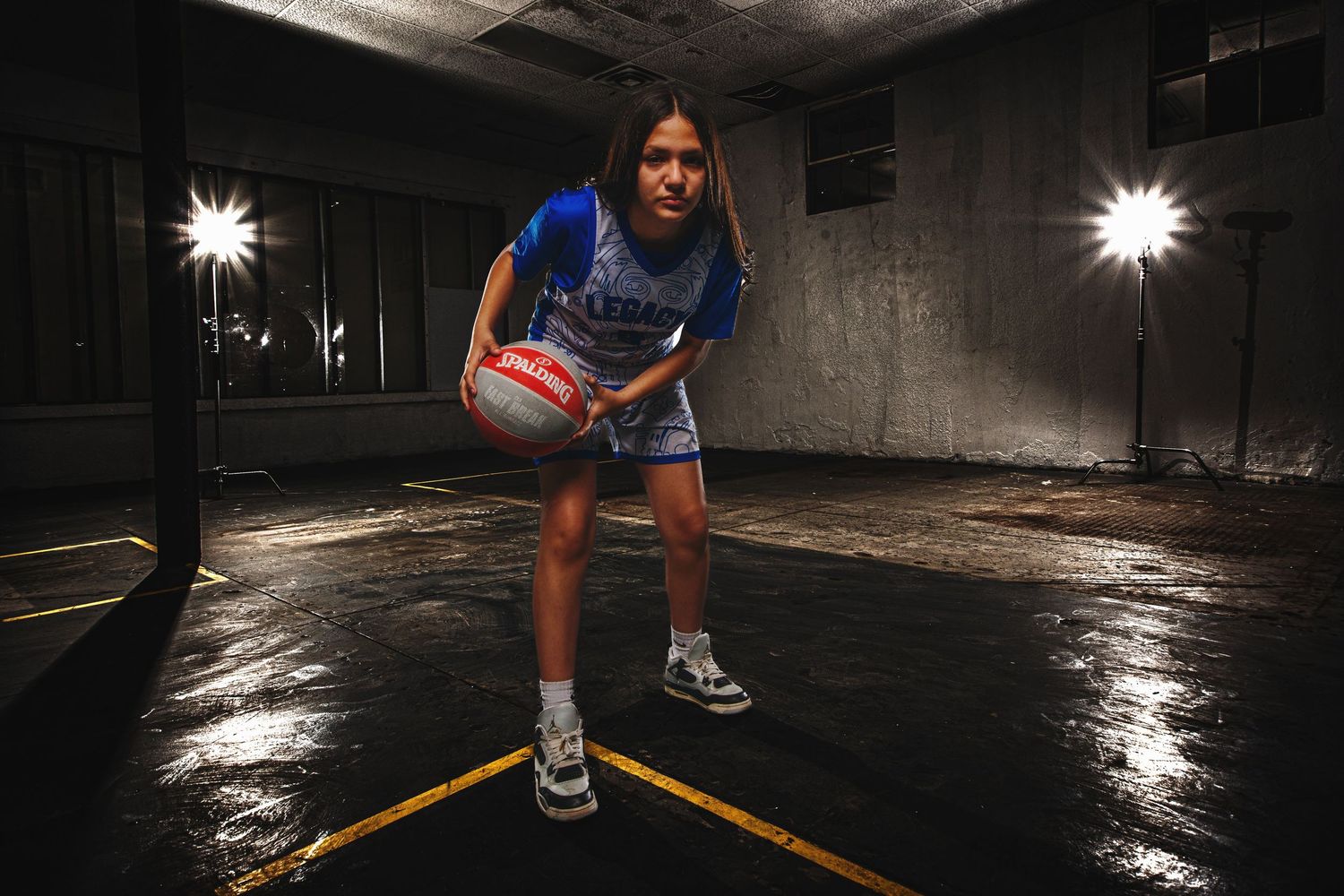 Young female basketball player in blue uniform dribbling ball in dramatic studio setting with bright lights.